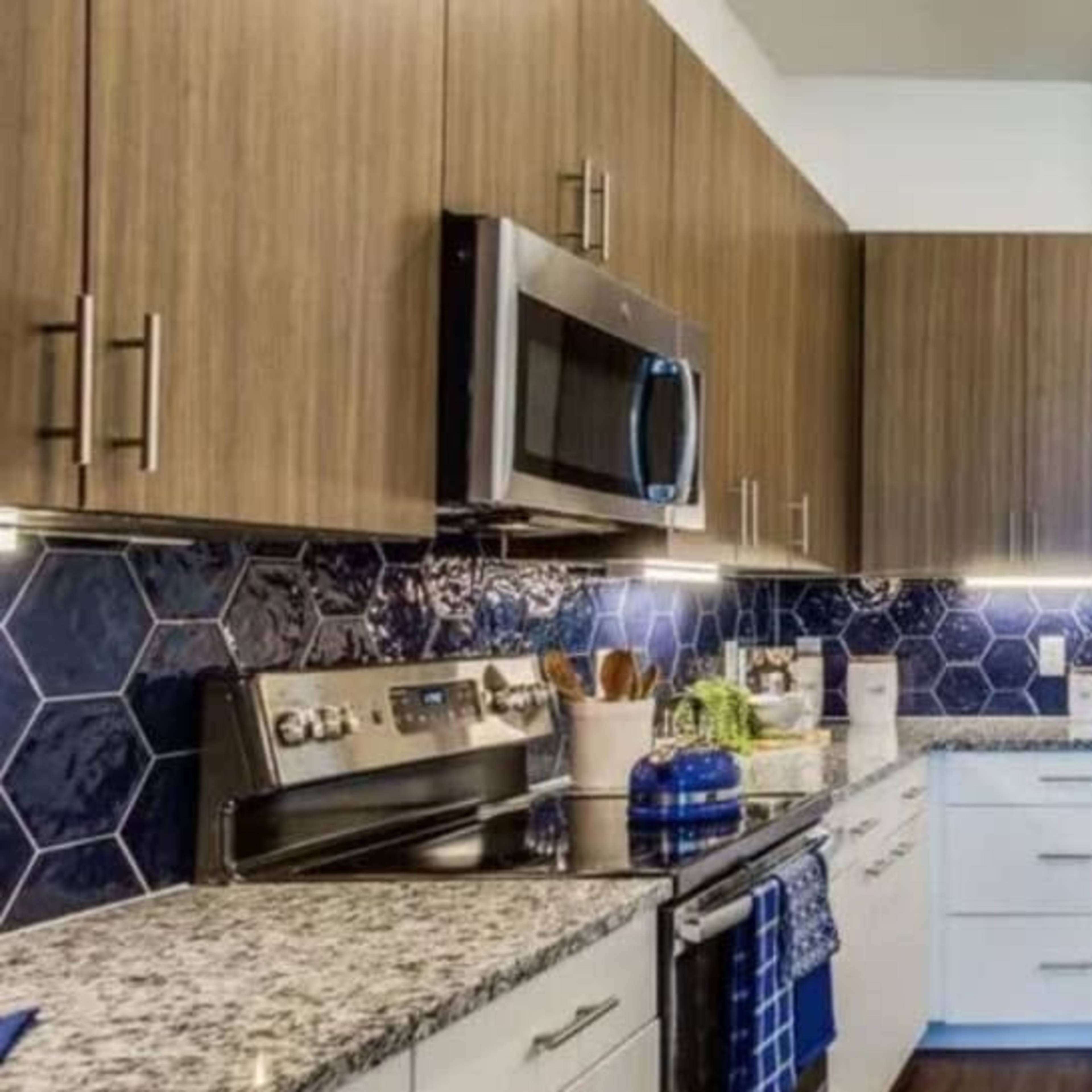 A modern kitchen featuring wood-panelled cabinets, granite countertops, stainless steel appliances, and a hexagonal tile backsplash.