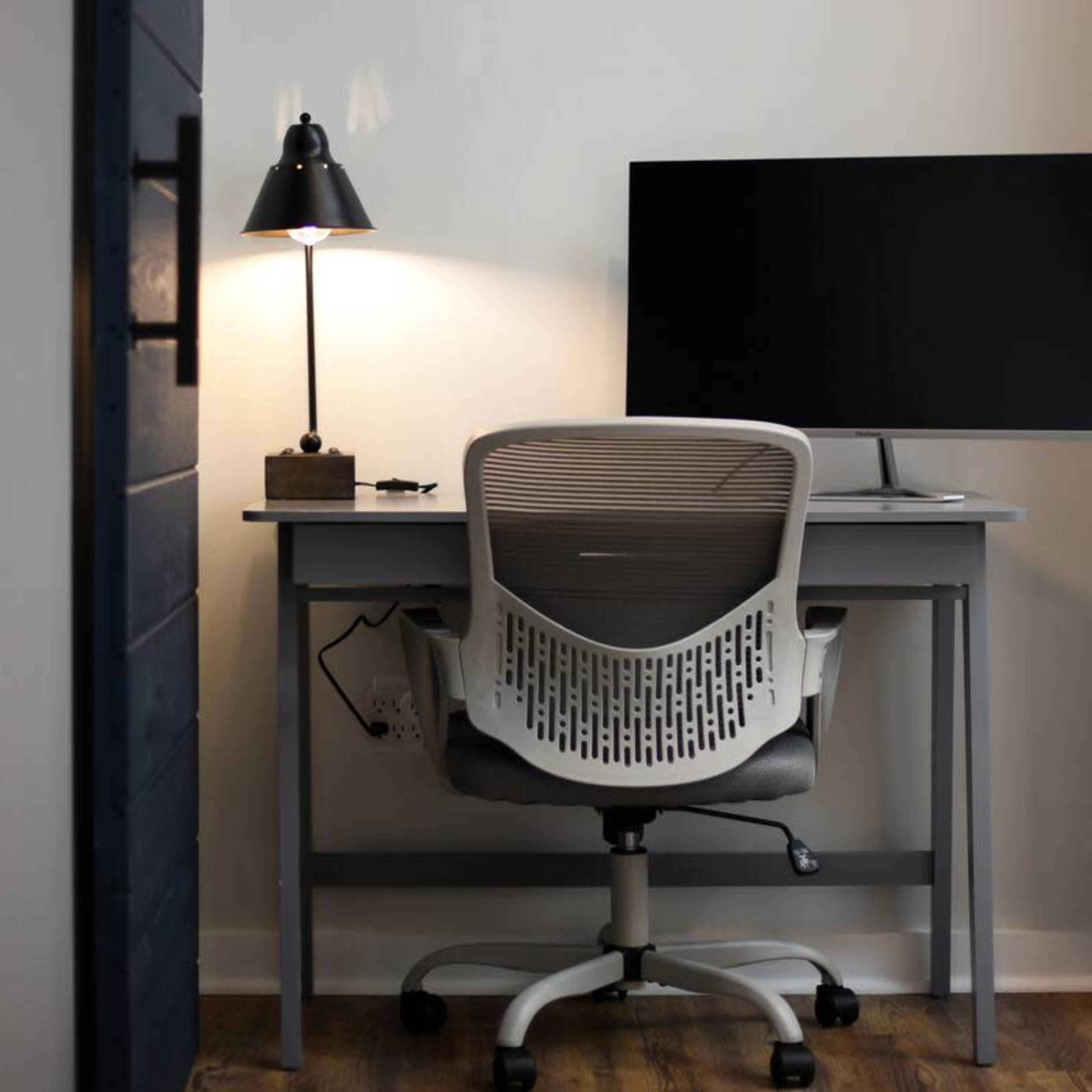 A modern workspace features a desk with a computer monitor, a desk lamp, and a comfortable office chair against a brick wall.