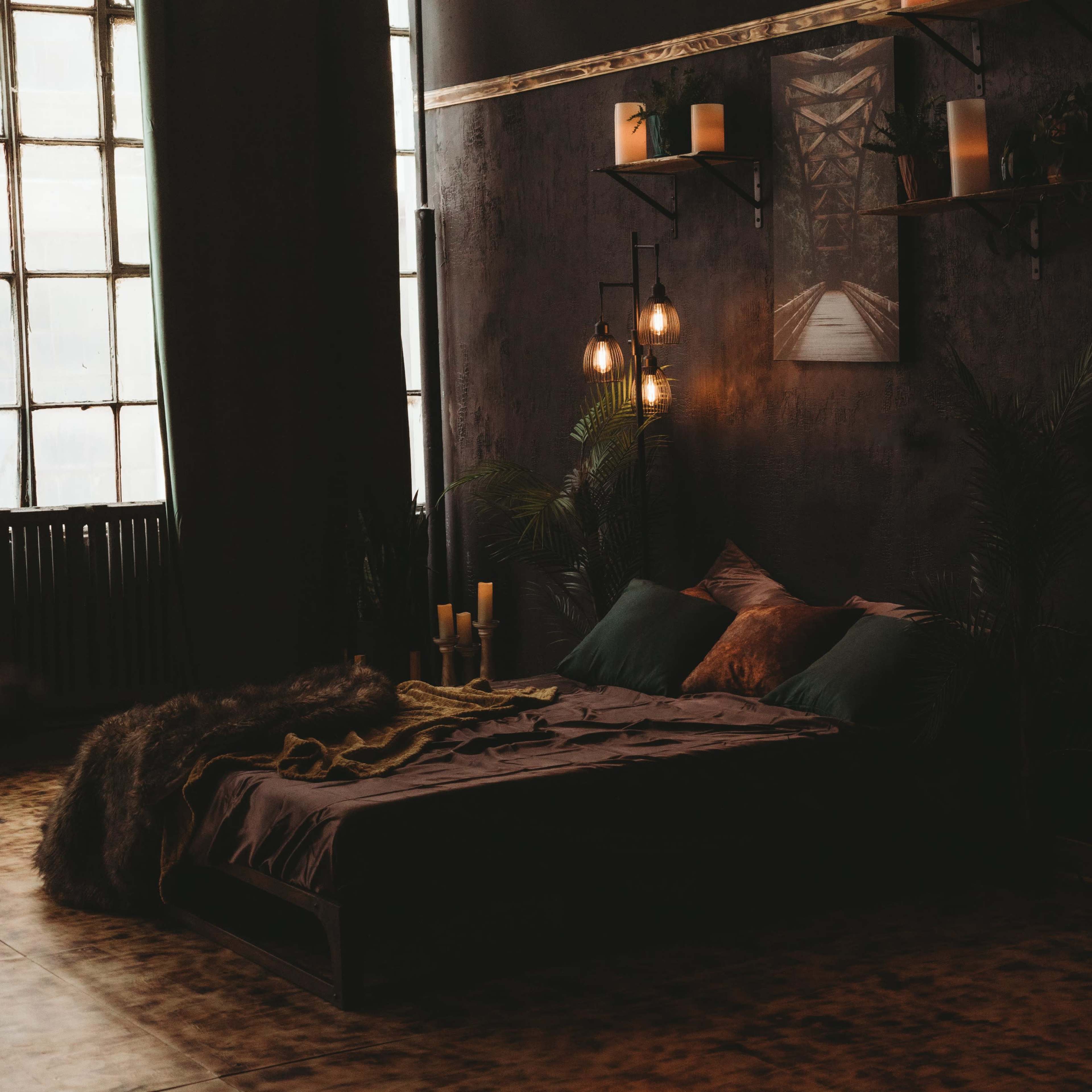 A dimly lit bedroom with a large bed, decorative plants, and candles arranged on shelves and surfaces.
