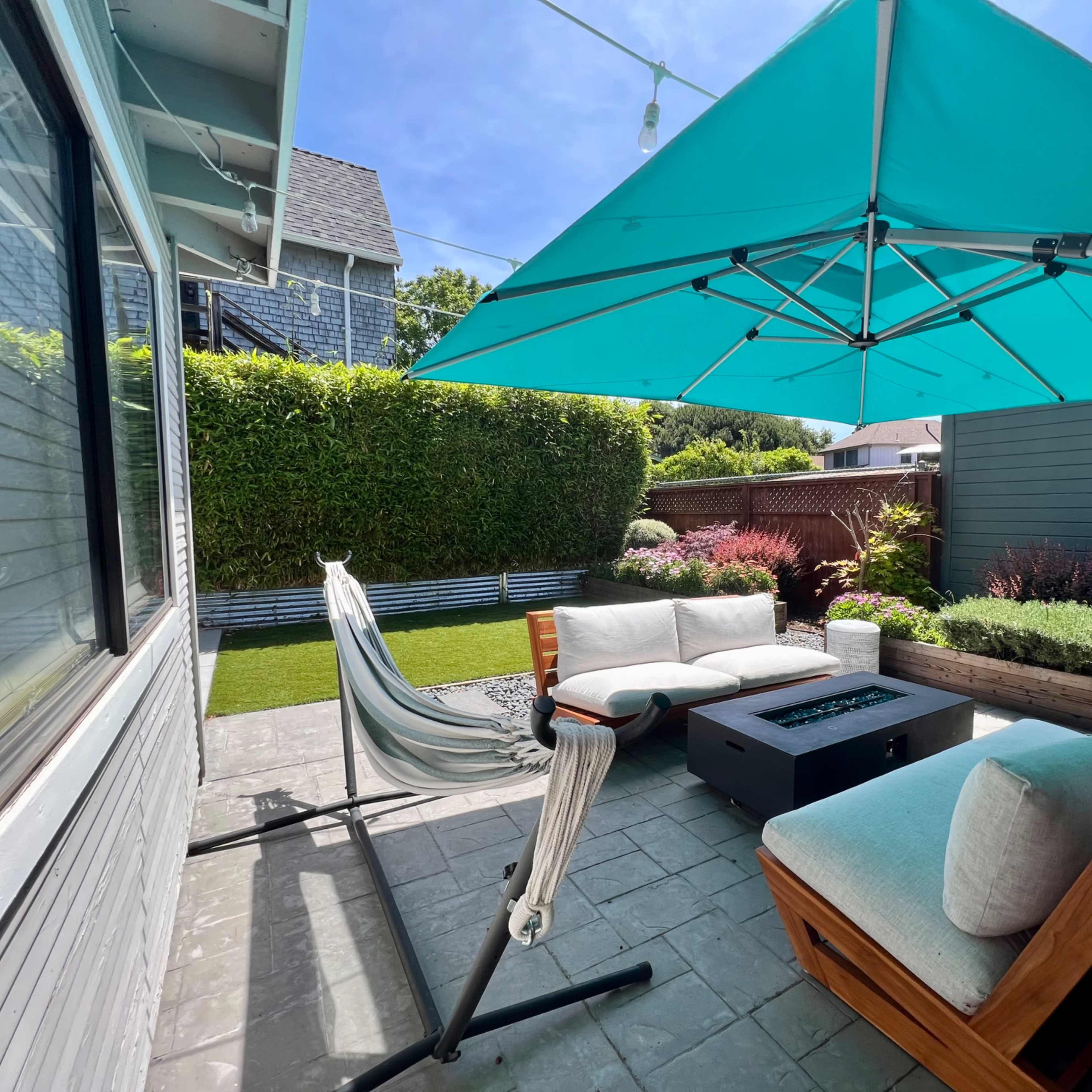 A modern outdoor patio featuring a teal canopy, comfortable seating, a fire table, and a hammock surrounded by greenery.