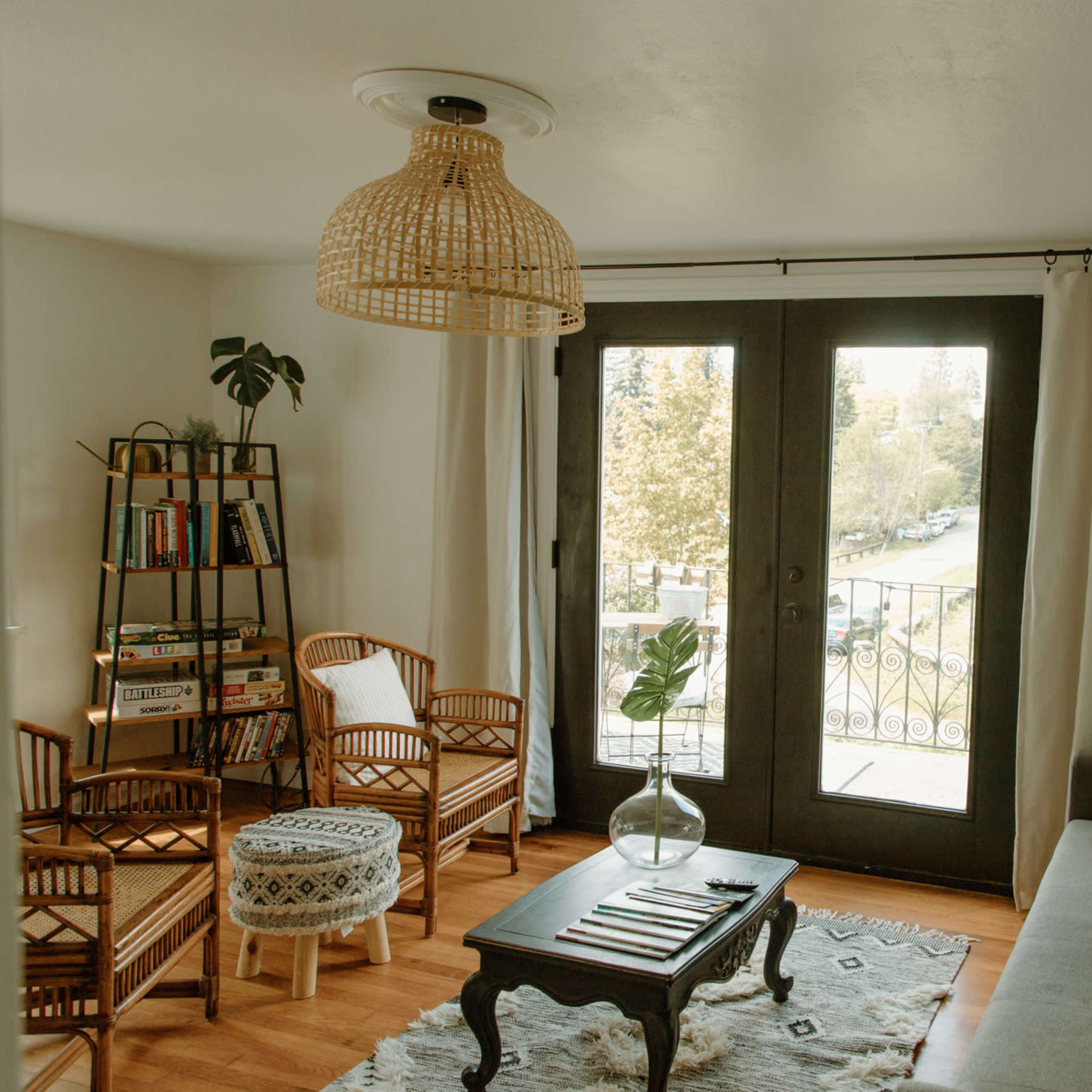A cozy living room with a gray sofa, rattan chairs, a wooden coffee table, and large doors that open to a balcony overlooking a tree-lined view.