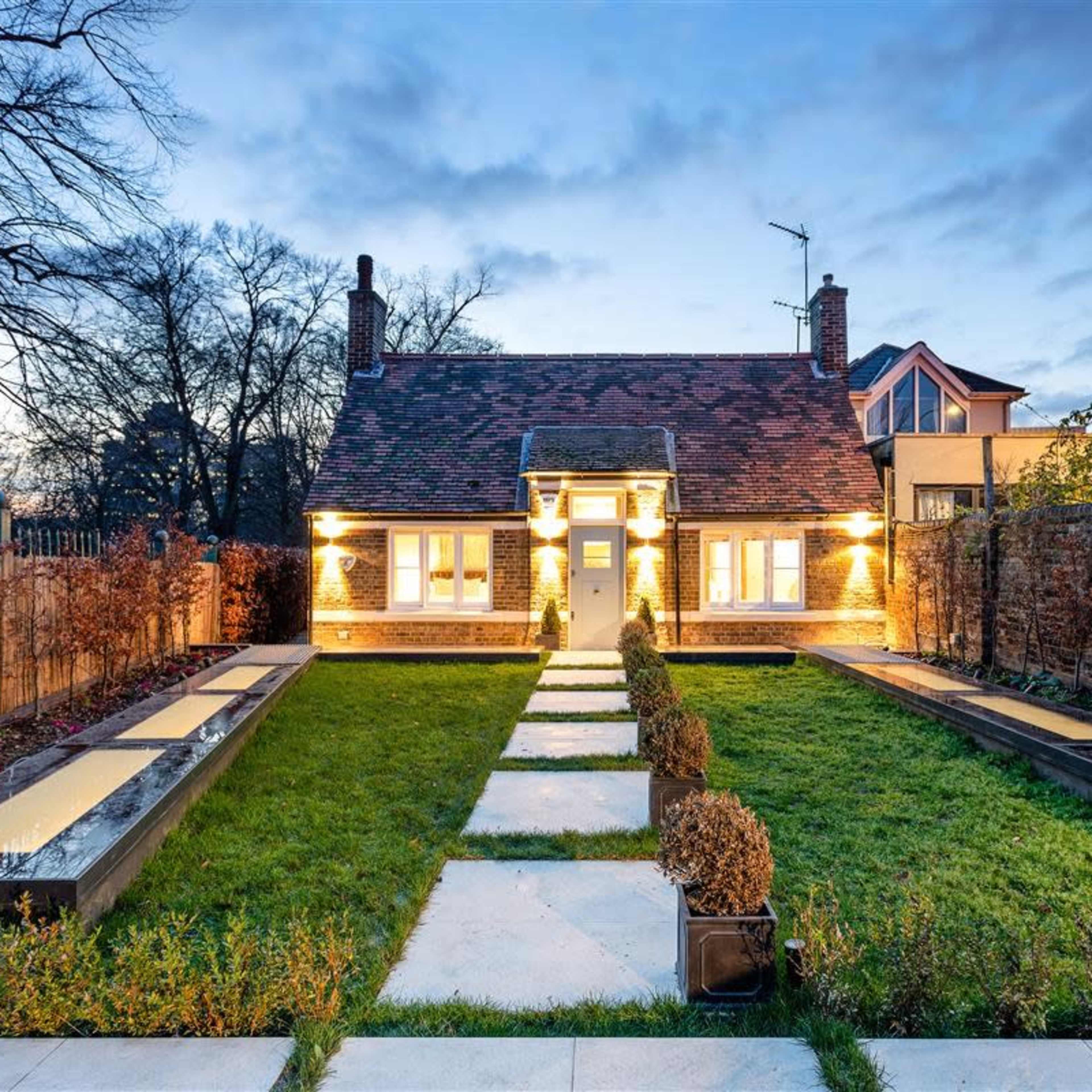 A well-lit house with a manicured lawn and walkways, surrounded by landscaped gardens and a stone wall.