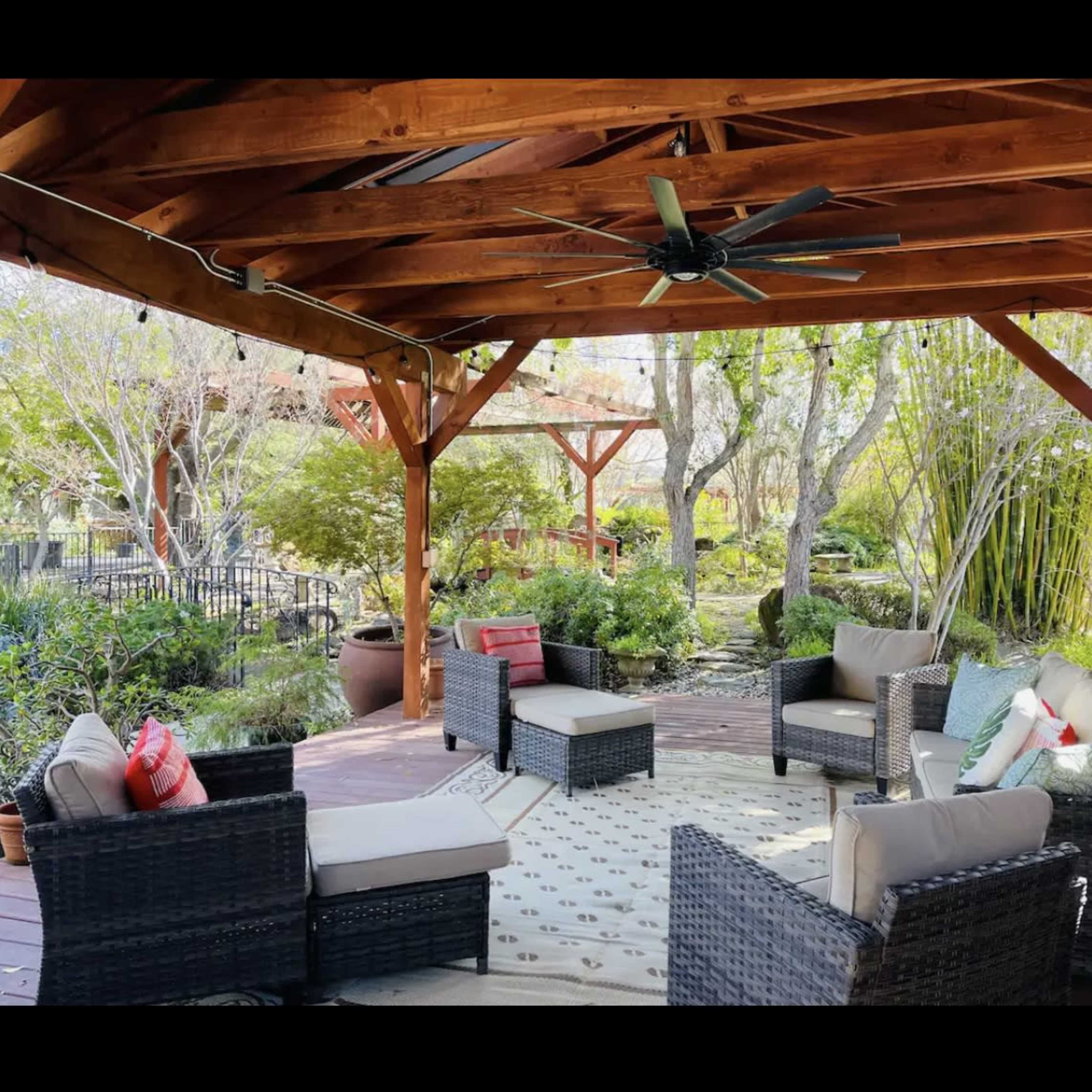 A covered outdoor seating area with wicker furniture arranged around a patterned rug in a landscaped garden.
