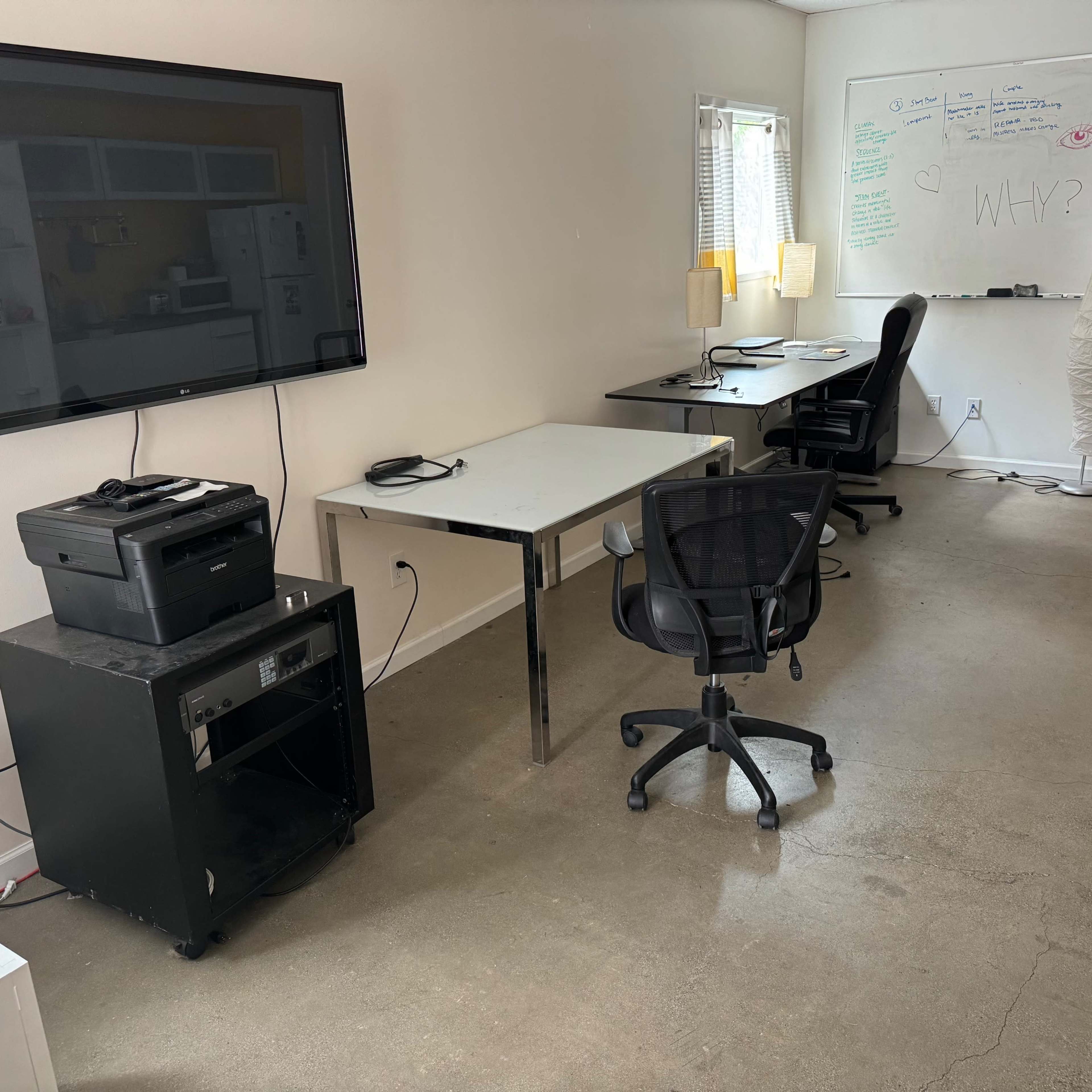 A simple office space featuring a large television on the wall, a printer on a black stand, a whiteboard with writing, and two workstations with chairs.