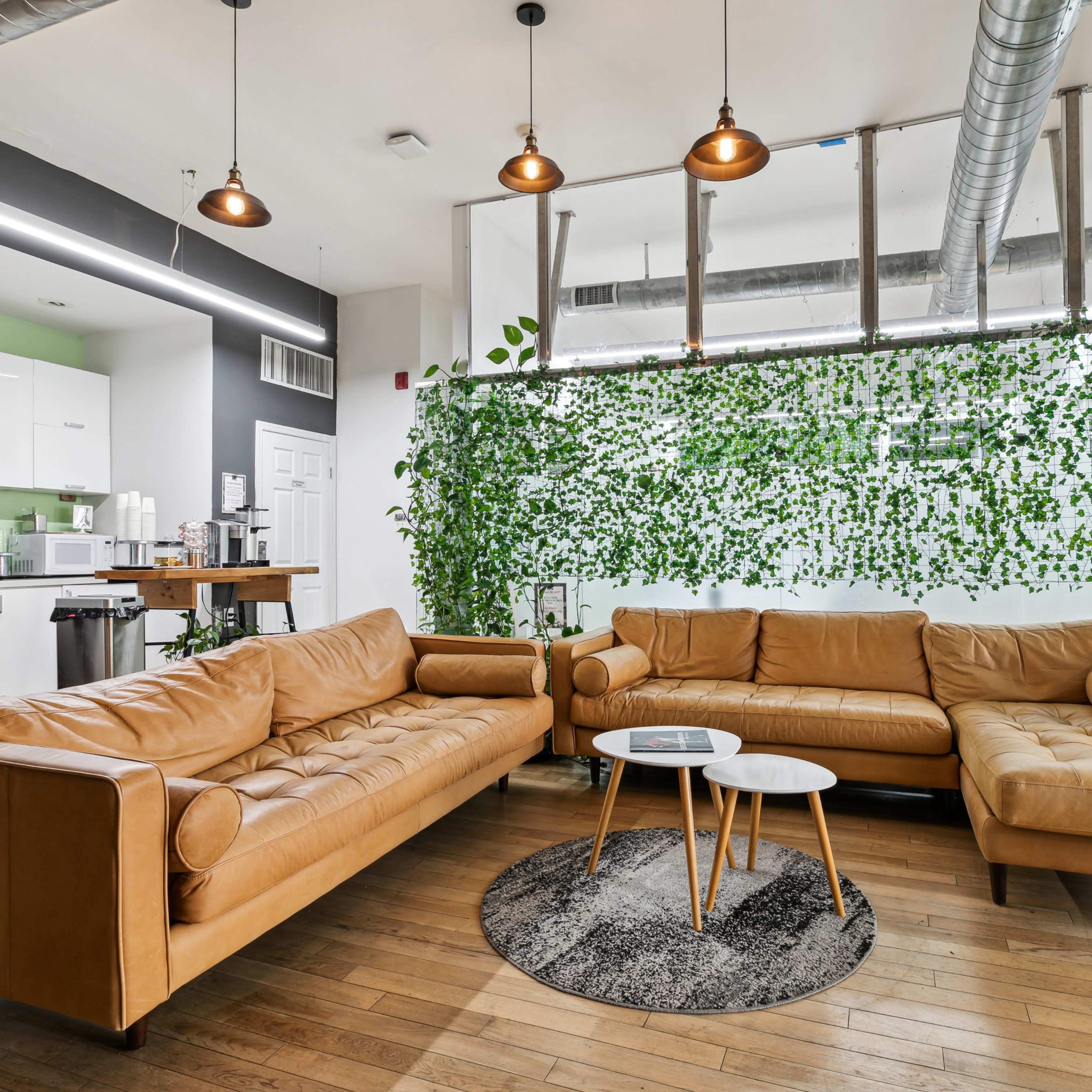A modern interior space featuring a large, tan leather sectional sofa, a round coffee table, and a wall with green foliage, with a kitchenette visible in the background.