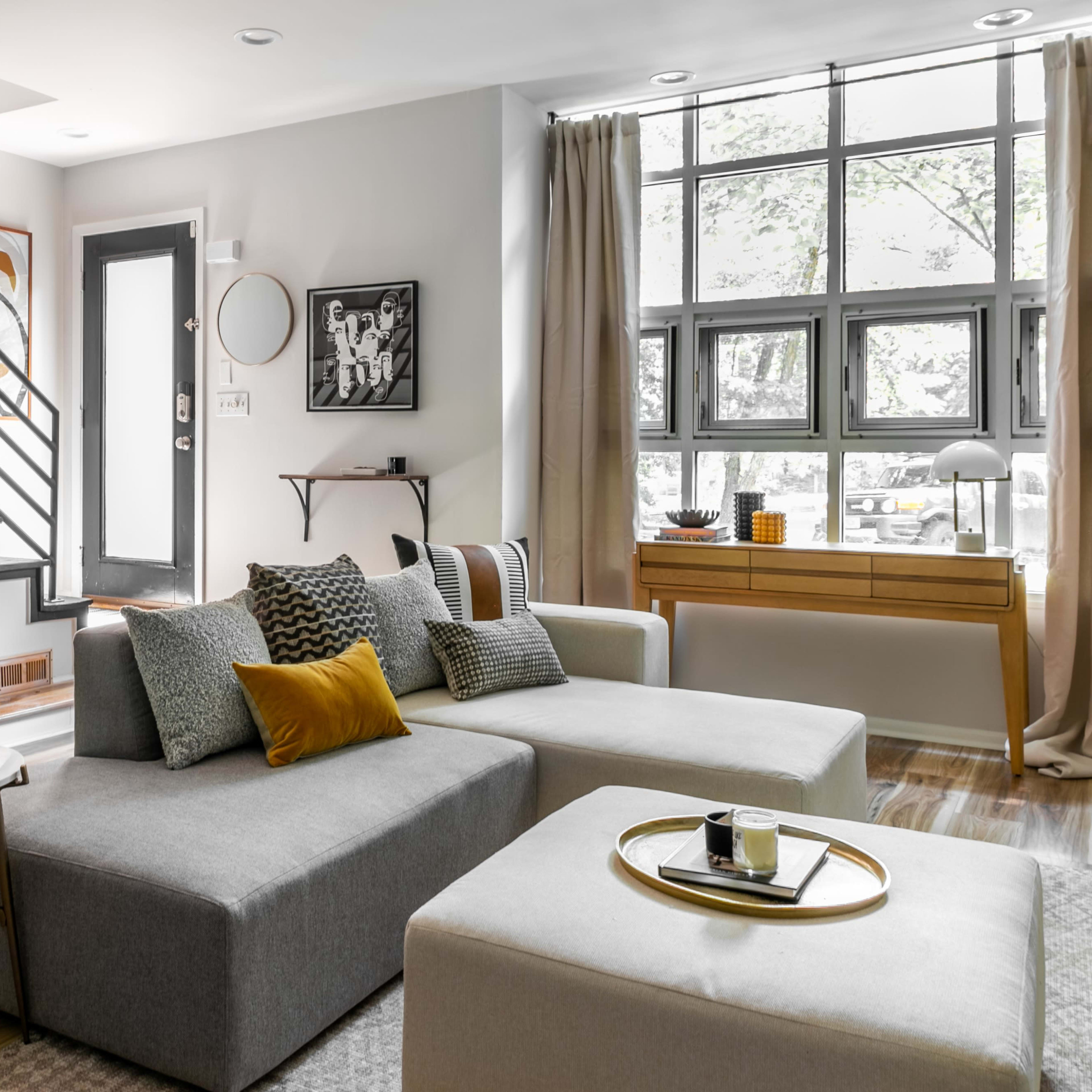 A modern living room with a gray sectional sofa, a coffee table, and a large window providing natural light, alongside a staircase in the background.