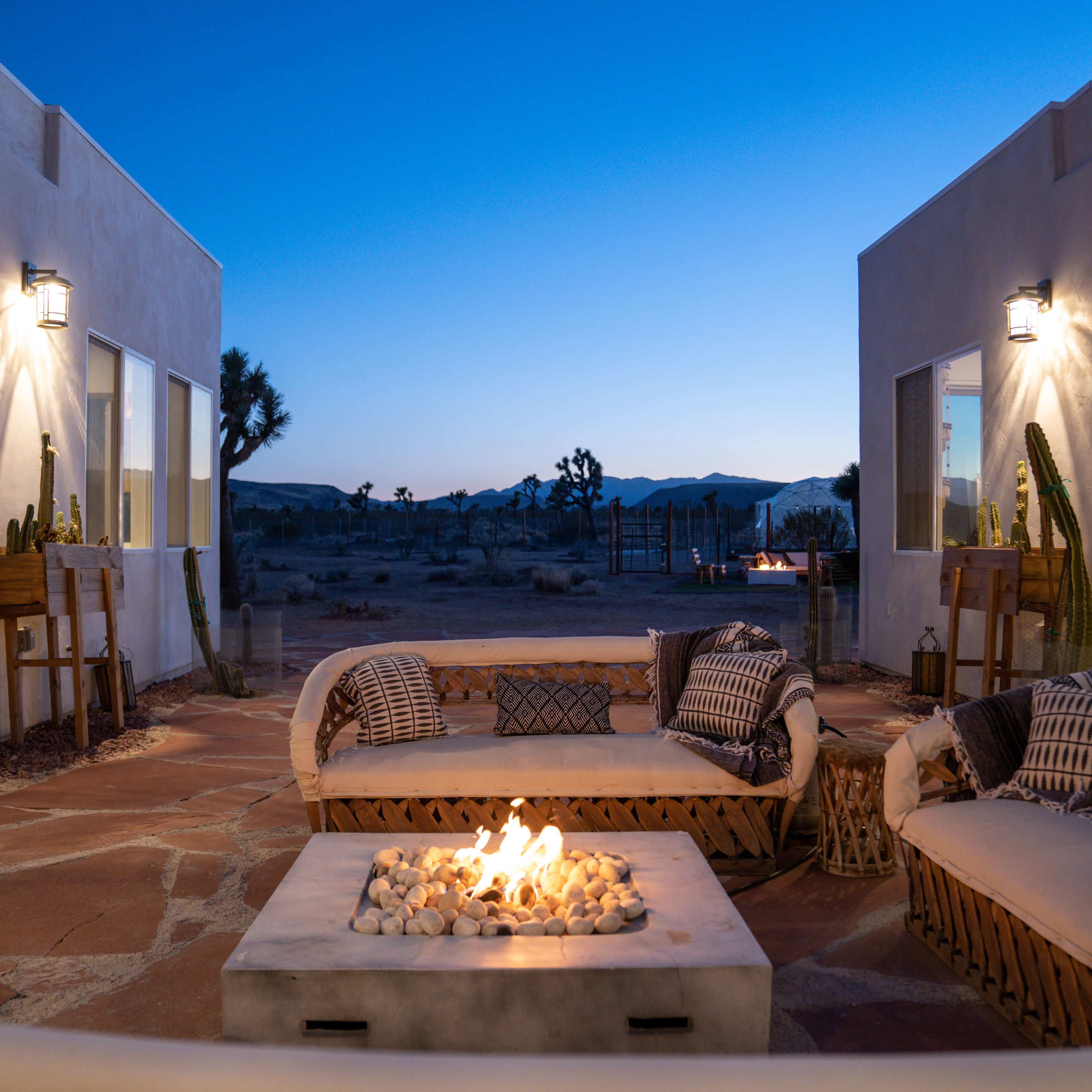 The scene shows an outdoor seating area with a fire pit surrounded by two adjacent buildings, set against a twilight sky with desert landscape in the background.