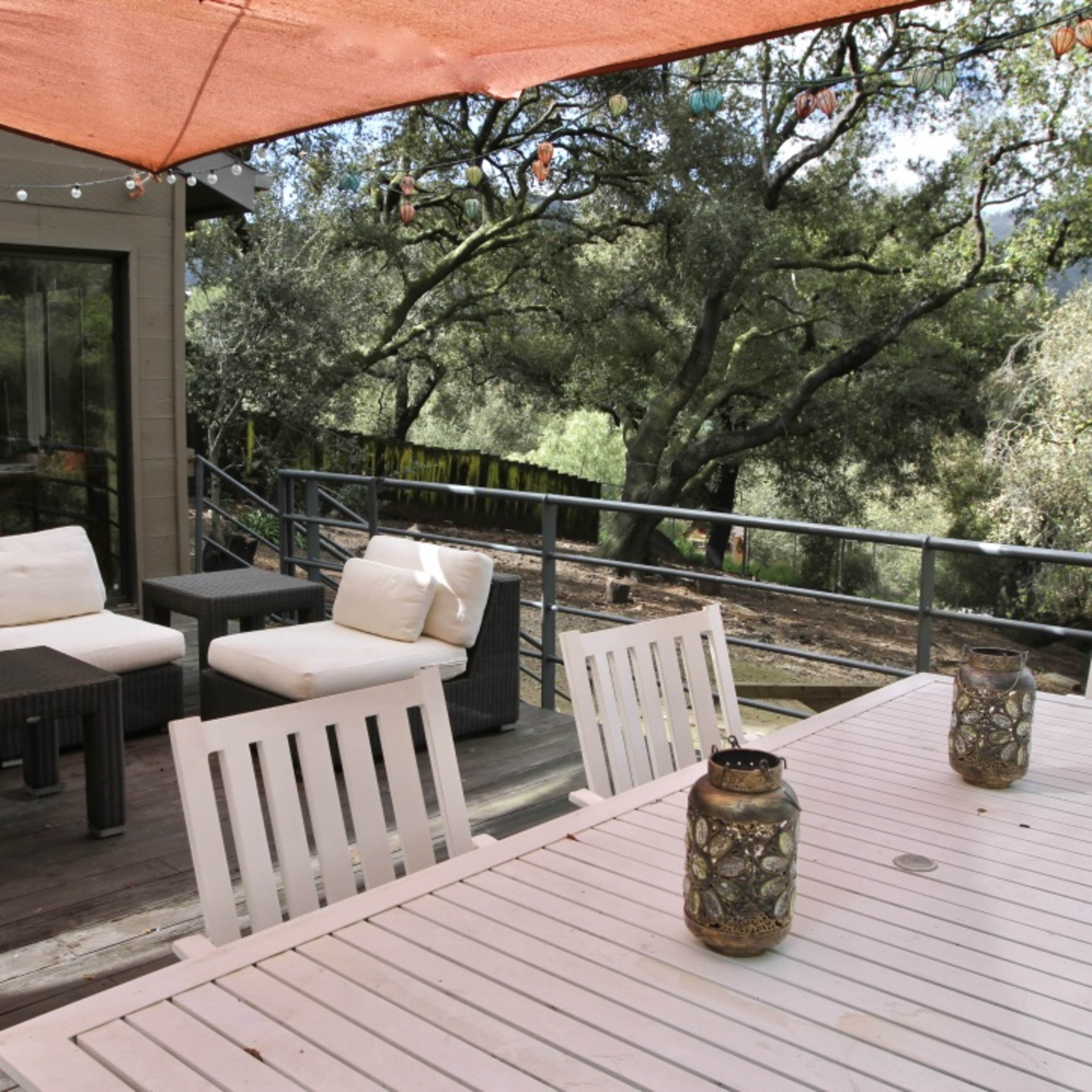 A wooden deck with a large table and seating area, shaded by an orange canopy, overlooks a green landscape with trees.