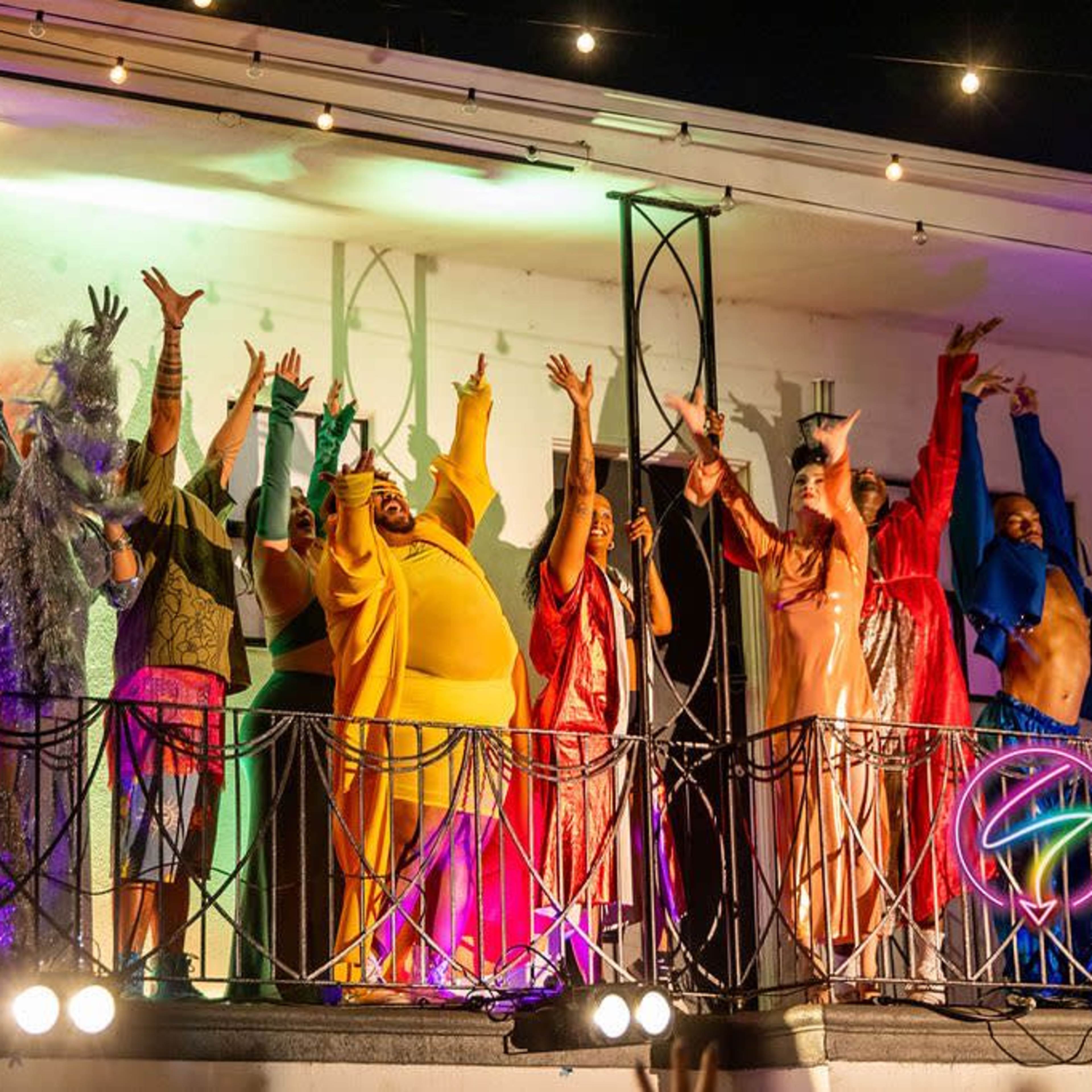 A group of performers dressed in vibrant costumes is celebrating on a balcony under colorful lights.