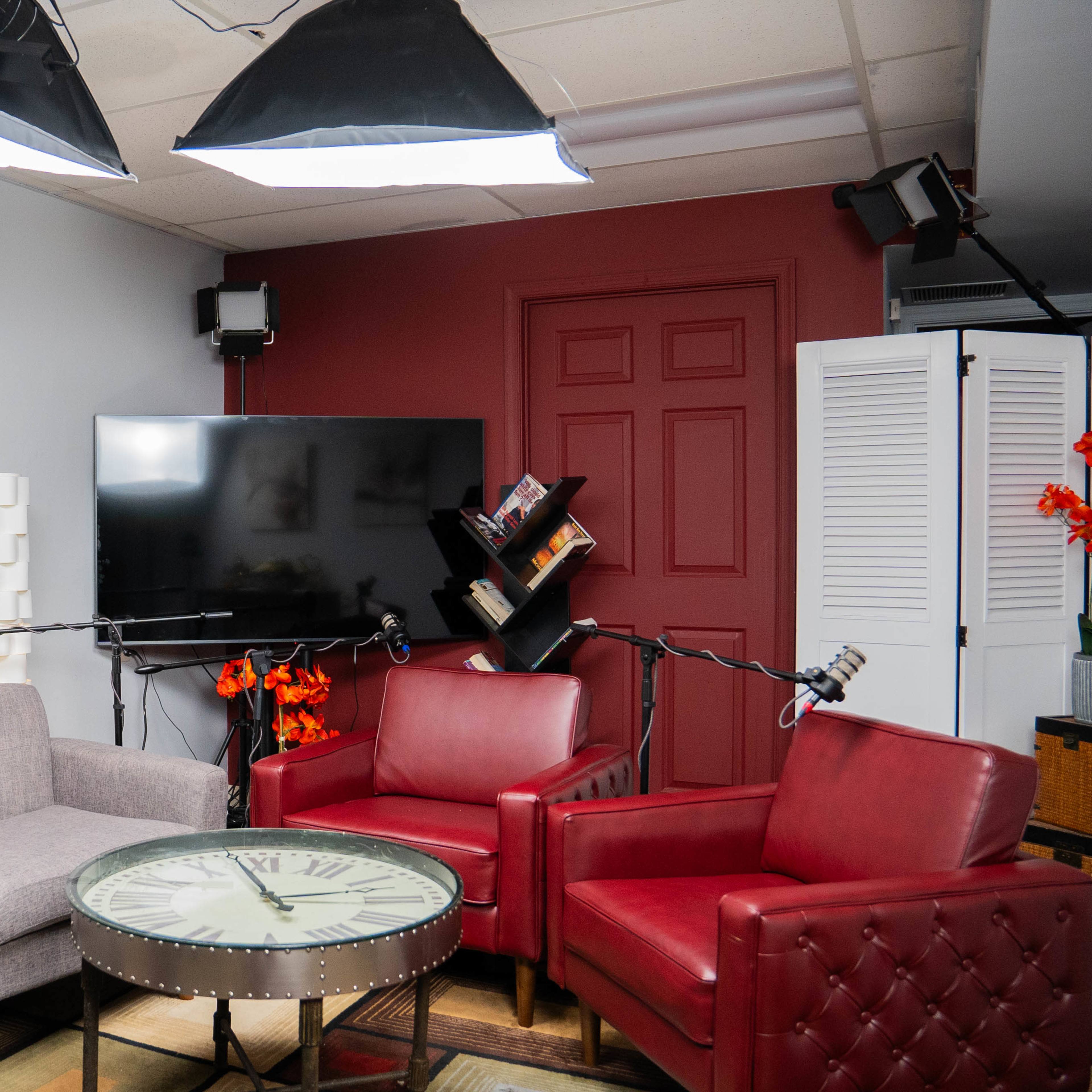 A modern studio space featuring a gray sofa, two red chairs, a round table with a clock design, and a large television on the wall, all illuminated by overhead lights.
