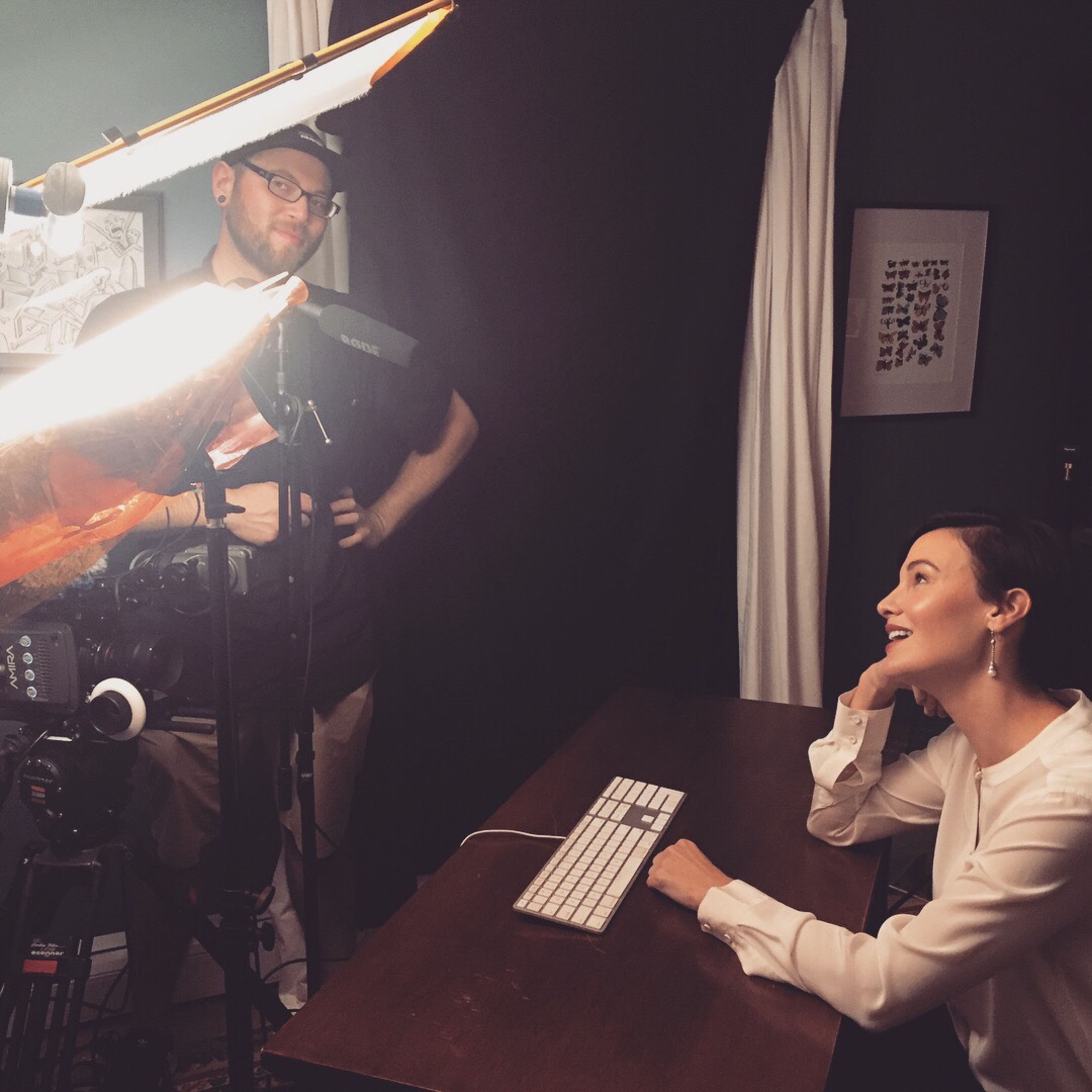 A woman sits at a desk with a keyboard while a man adjusts lighting equipment behind her in a dimly lit room.