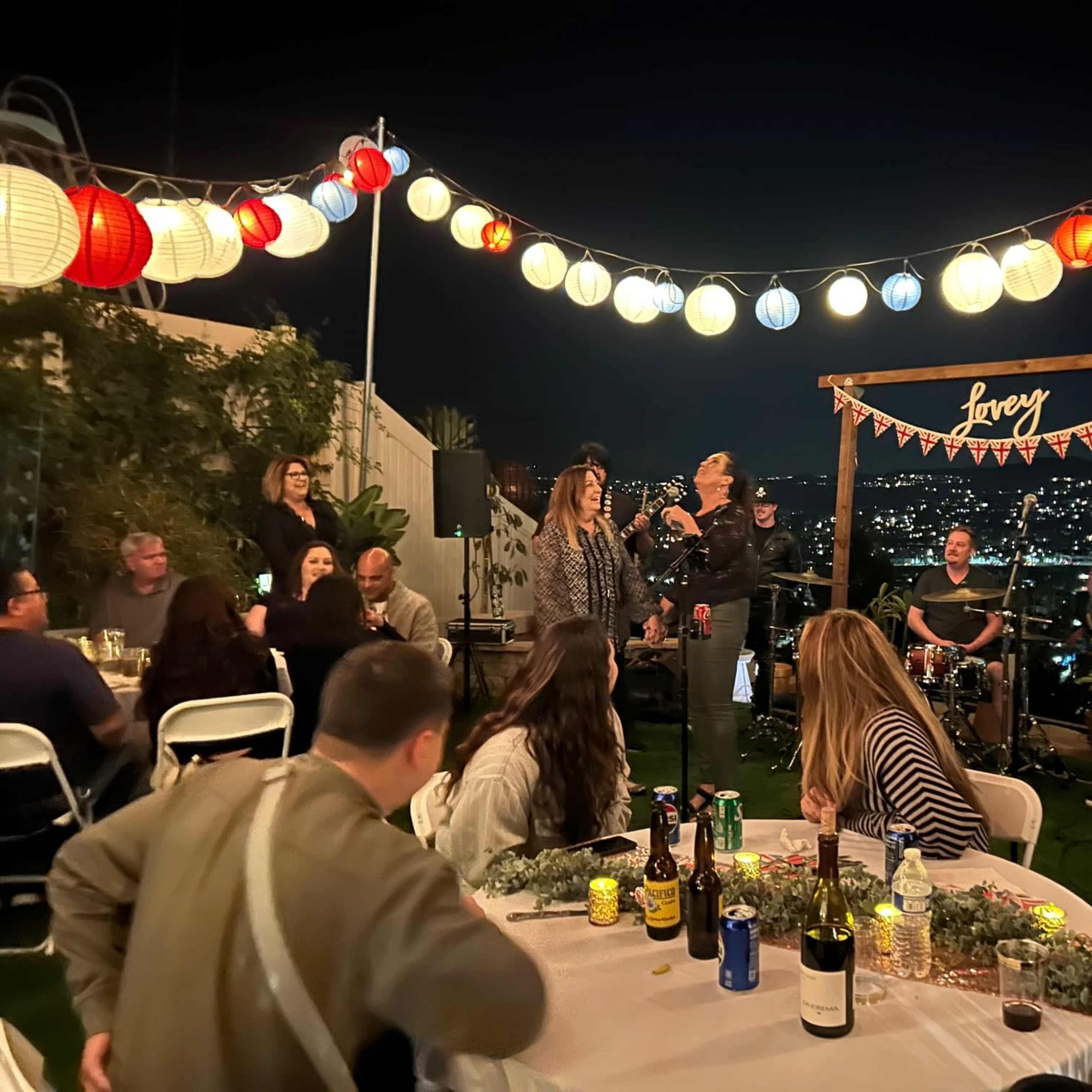 A rooftop party features guests gathered around tables with drinks, while performers entertain in front of city views illuminated by decorative lanterns.