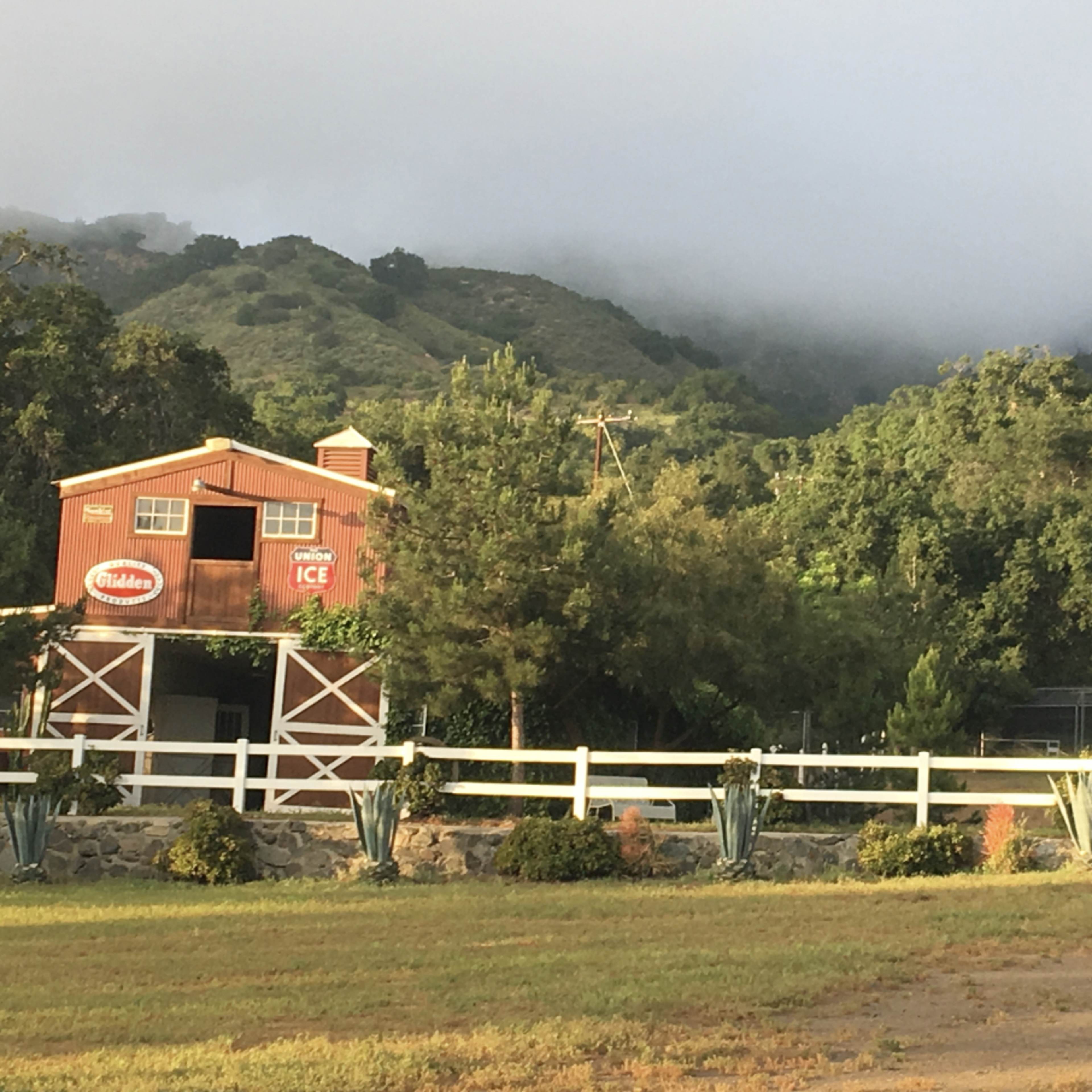 A red barn with a windmill and an old tractor is set against a backdrop of rolling green hills and low-hanging fog.