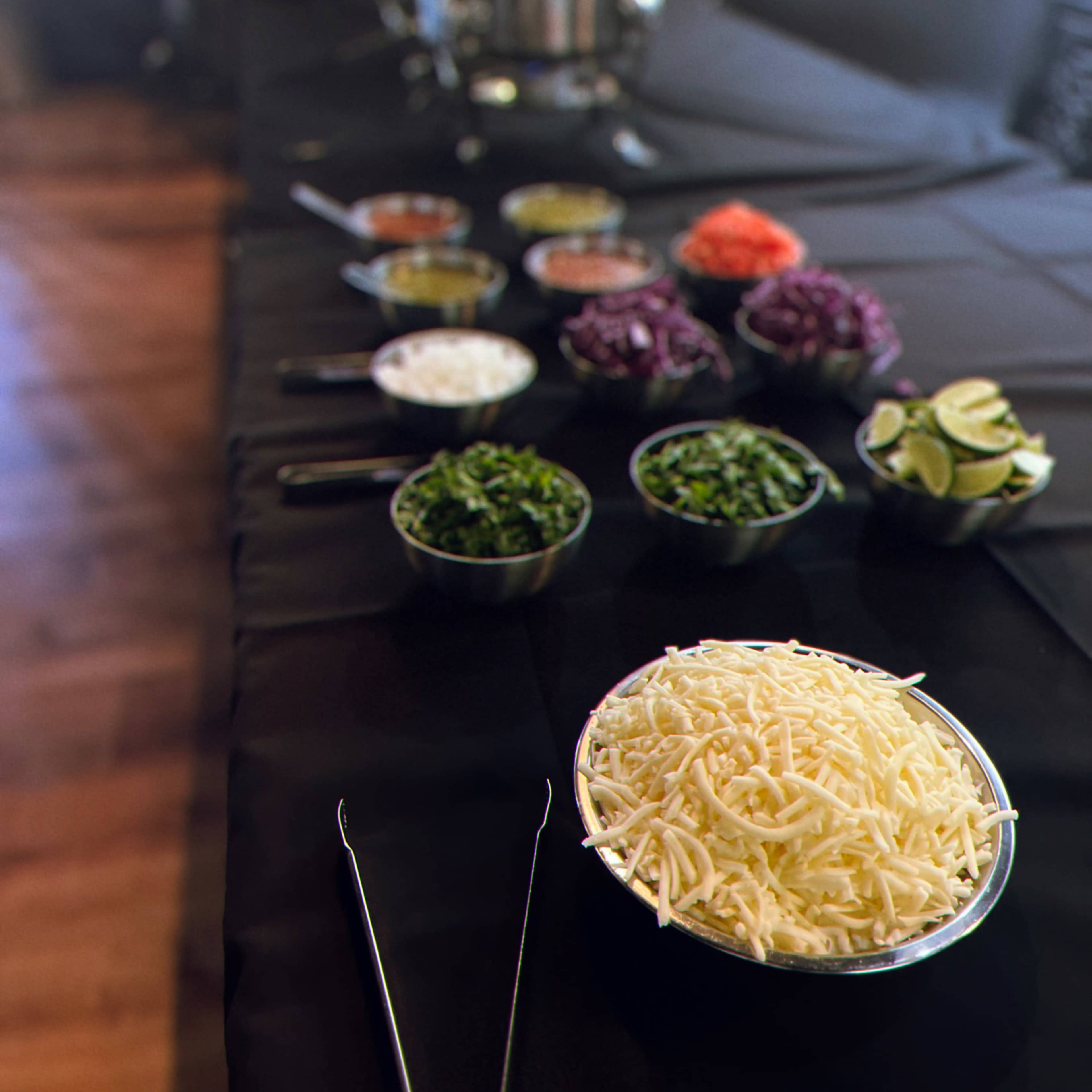 A long table is set with various bowls of toppings, including shredded cheese, chopped vegetables, and garnishes, arranged alongside serving utensils and containers.