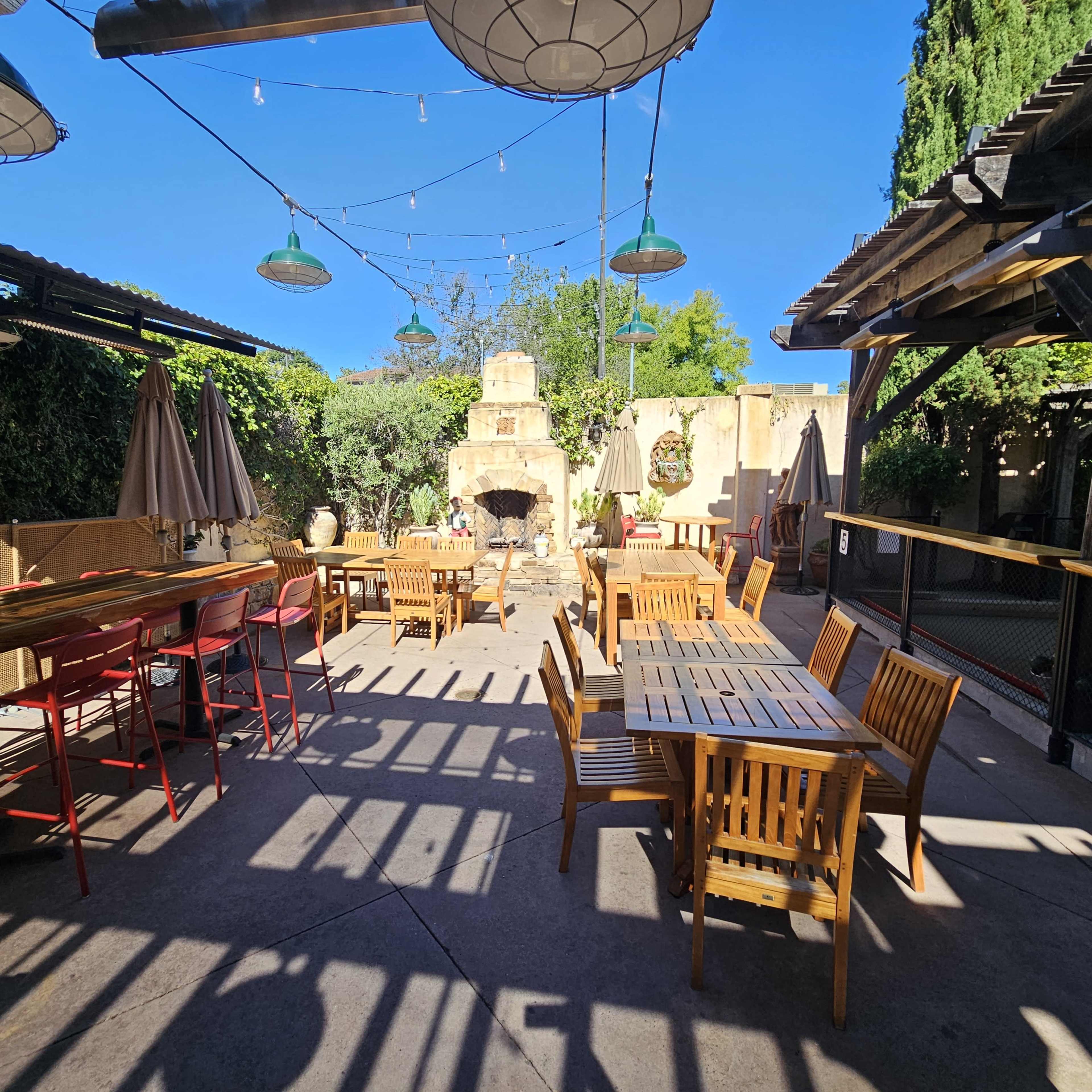 An outdoor seating area with wooden tables and chairs, a stone fireplace, and string lights overhead.