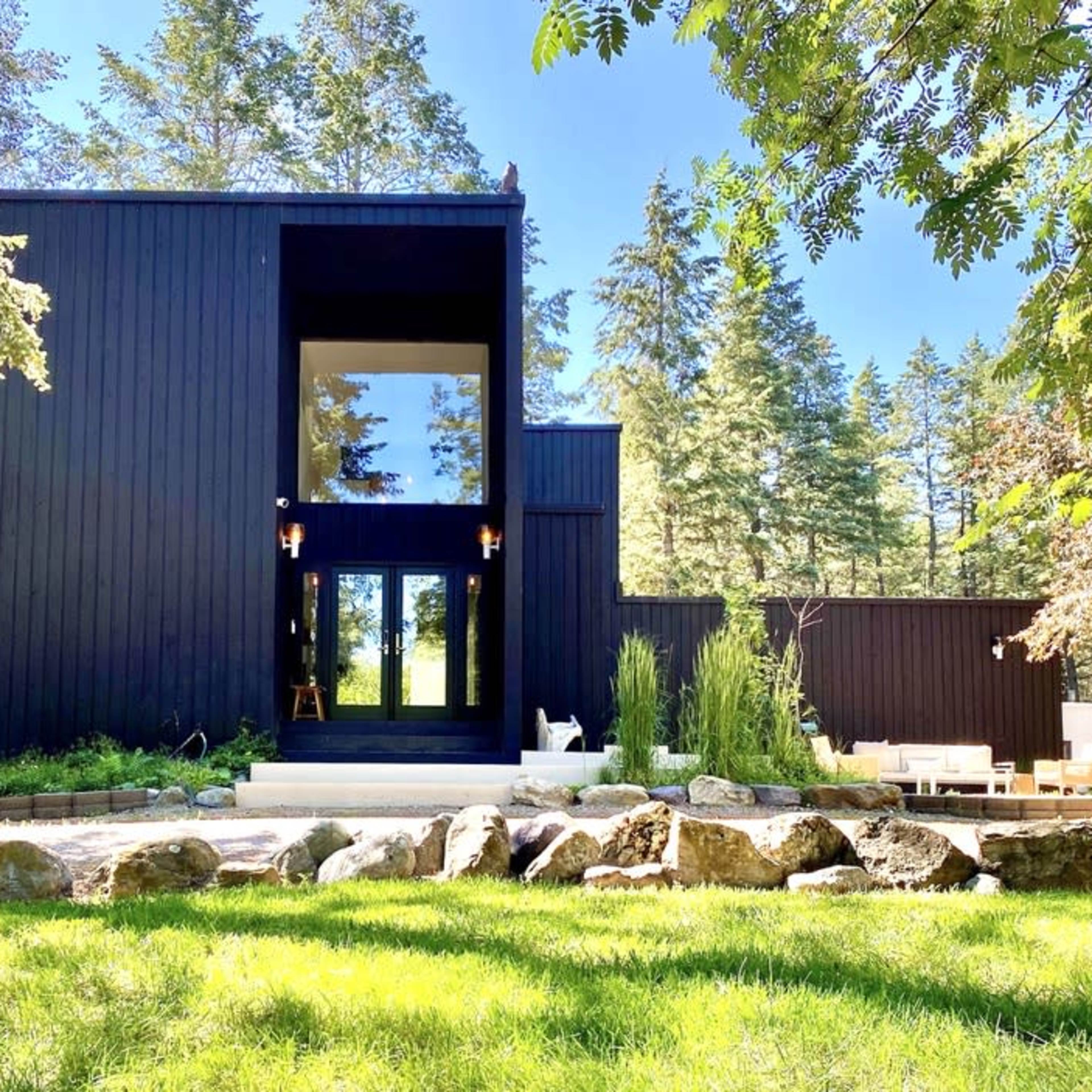 A modern black house with large windows is surrounded by green grass, trees, and landscaped rocks.
