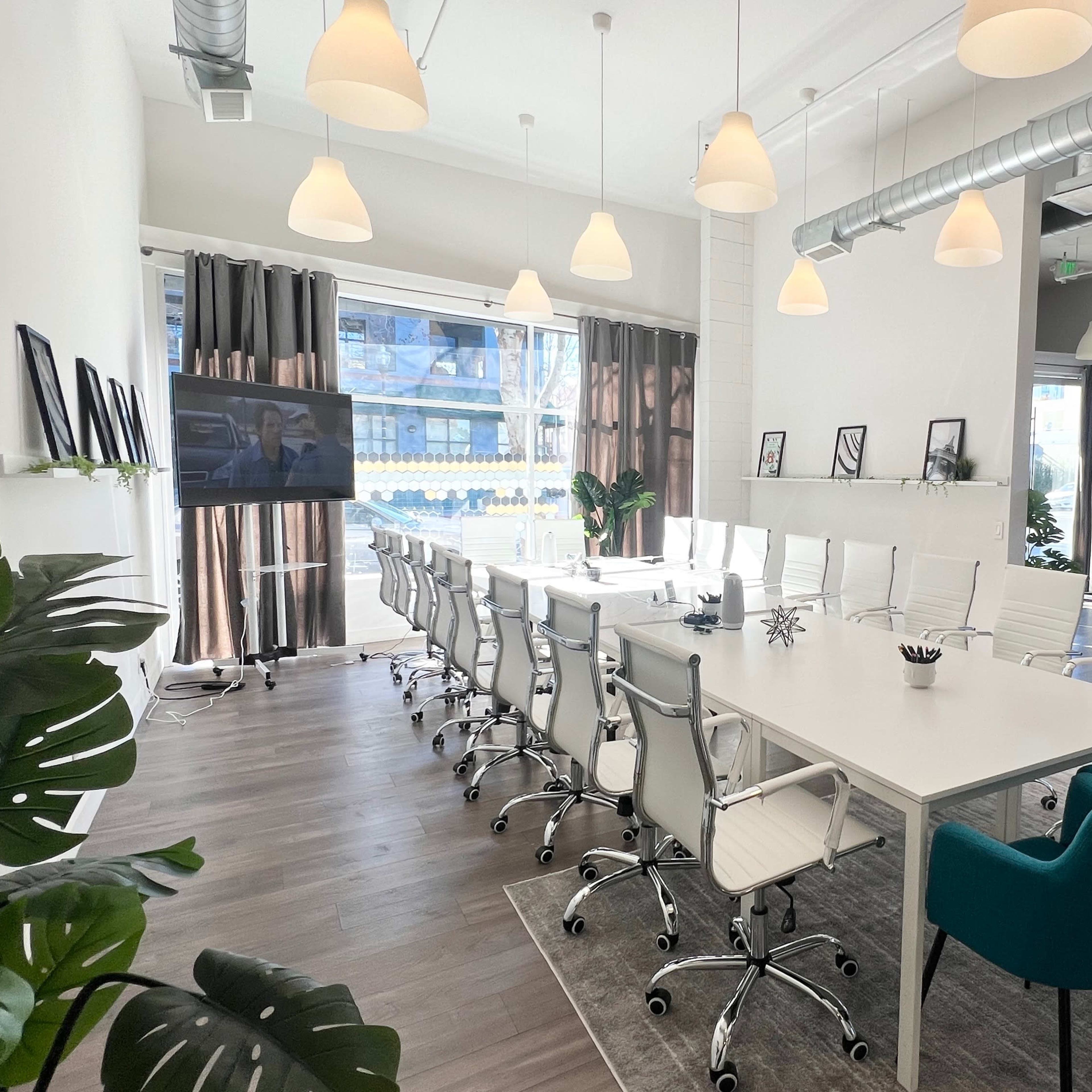 A modern conference room features a long white table surrounded by ergonomic chairs, with large windows and decorative plants.