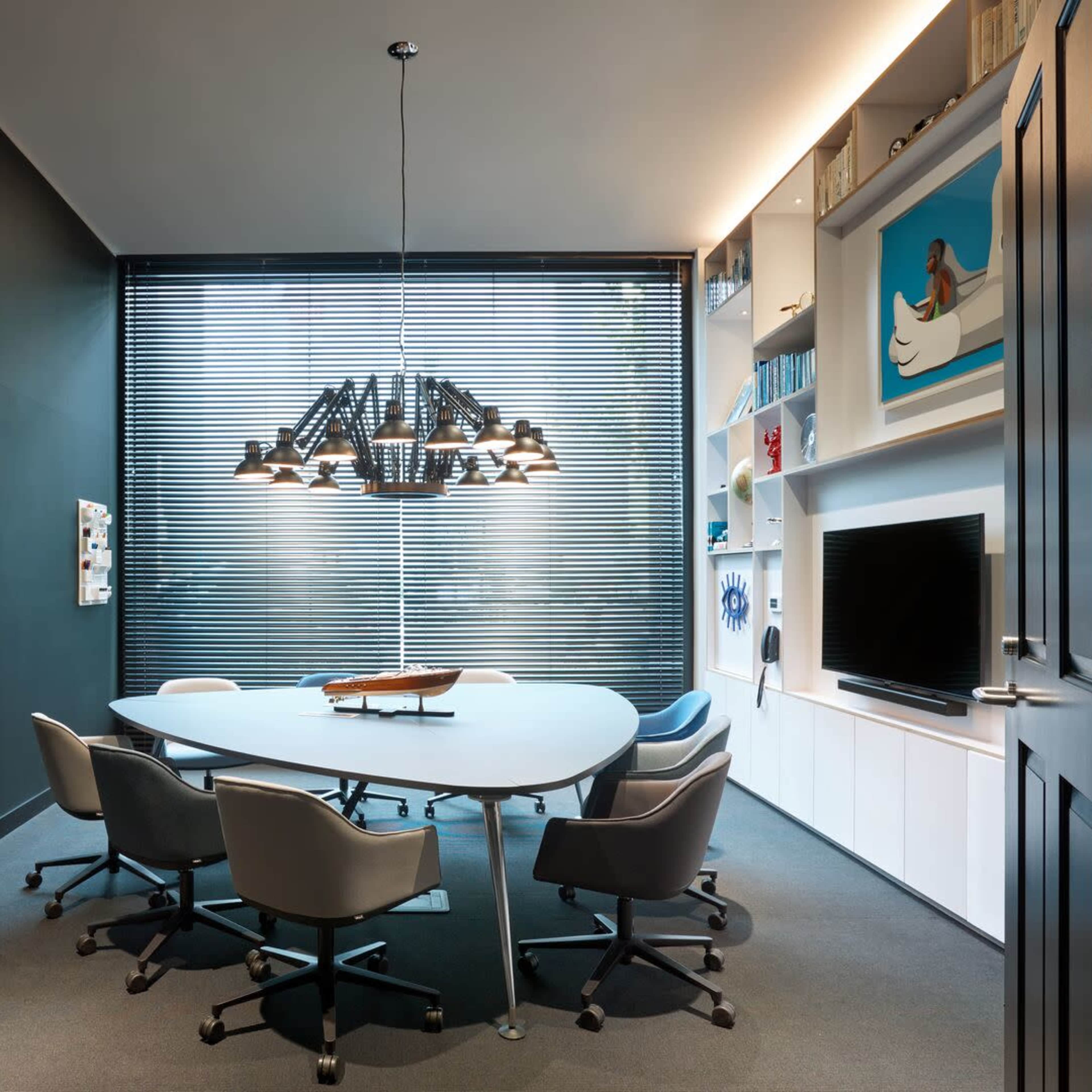 A modern conference room features a round table surrounded by six chairs, with a large pendant light overhead and shelves displaying decor, set against dark walls and window blinds.
