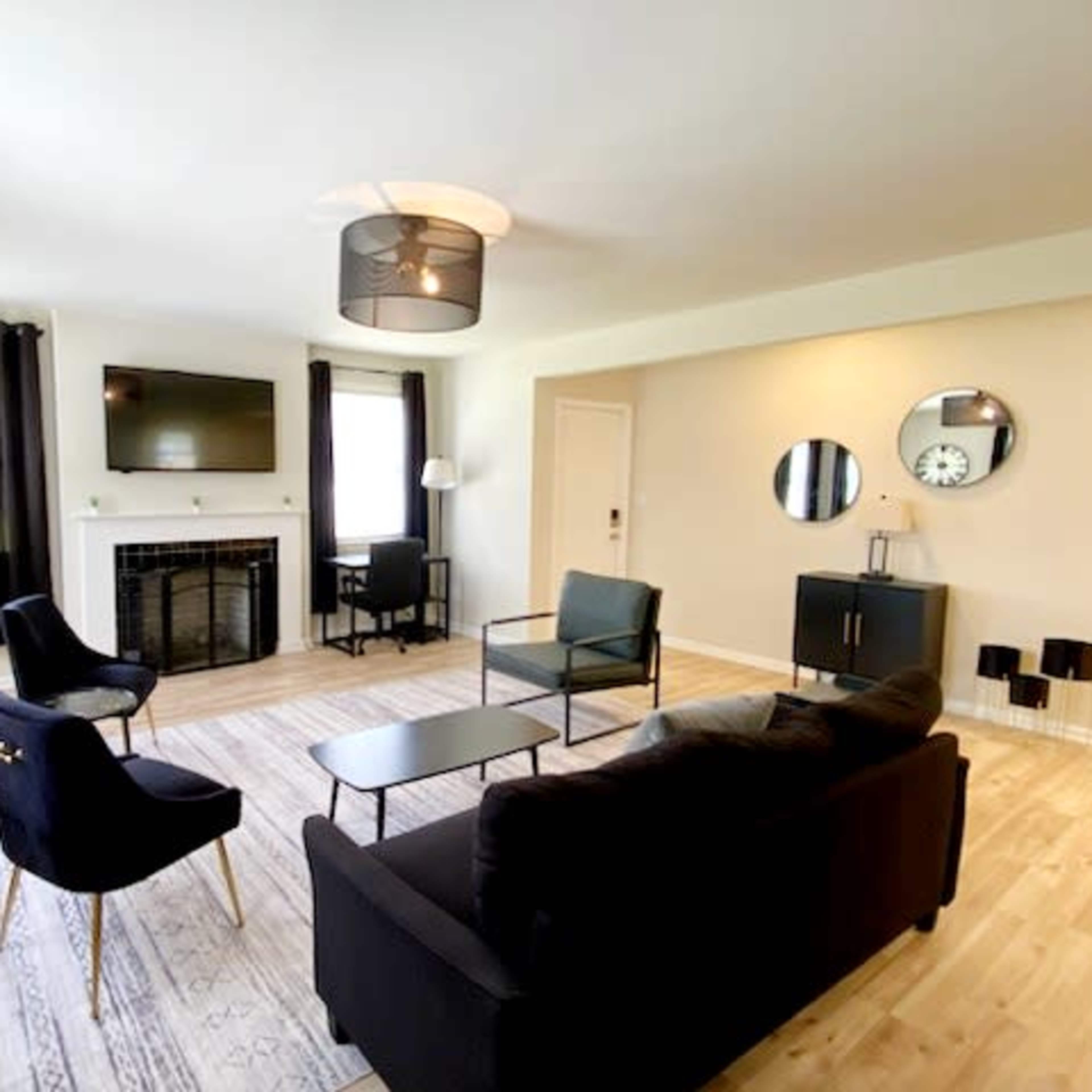 The living room features a central black sofa, two chairs, a coffee table, a fireplace, and a flat-screen TV mounted on the wall, with large windows dressed in dark curtains.