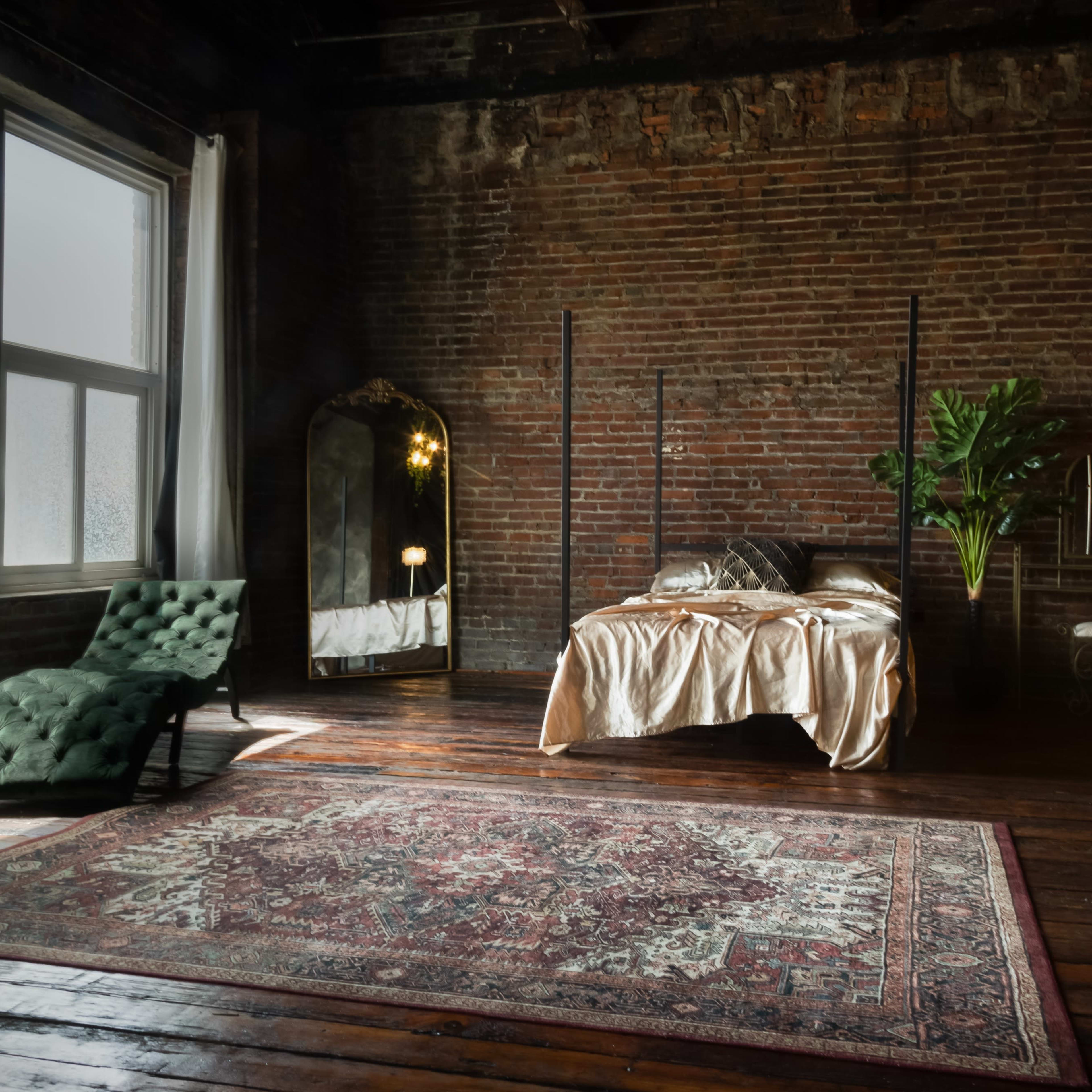 A bedroom with a vintage bed, a large mirror, a green lounge chair, and a potted plant in a room with exposed brick walls and wooden flooring.