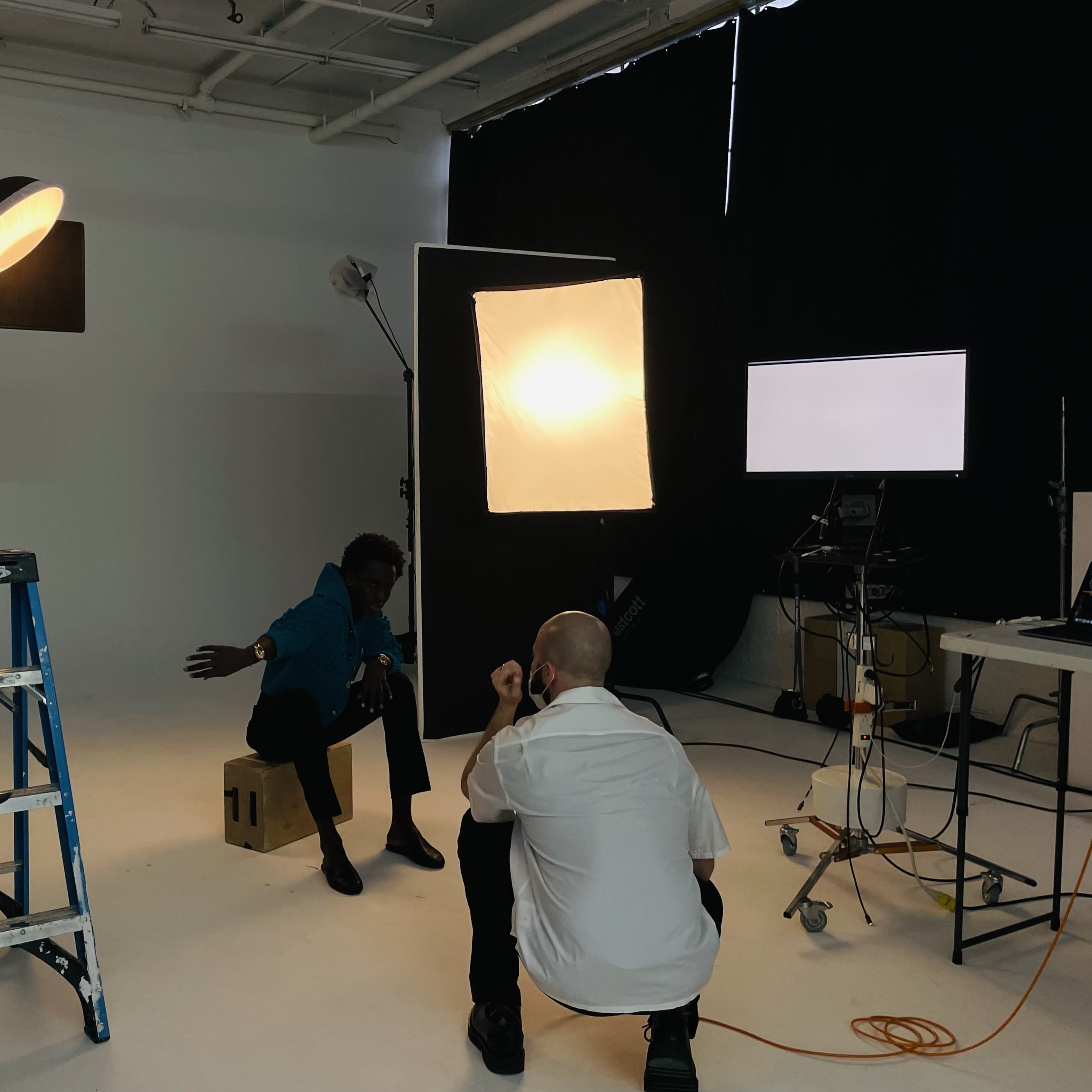 A photographer is directing a model sitting on a box in a studio illuminated by multiple lighting setups.