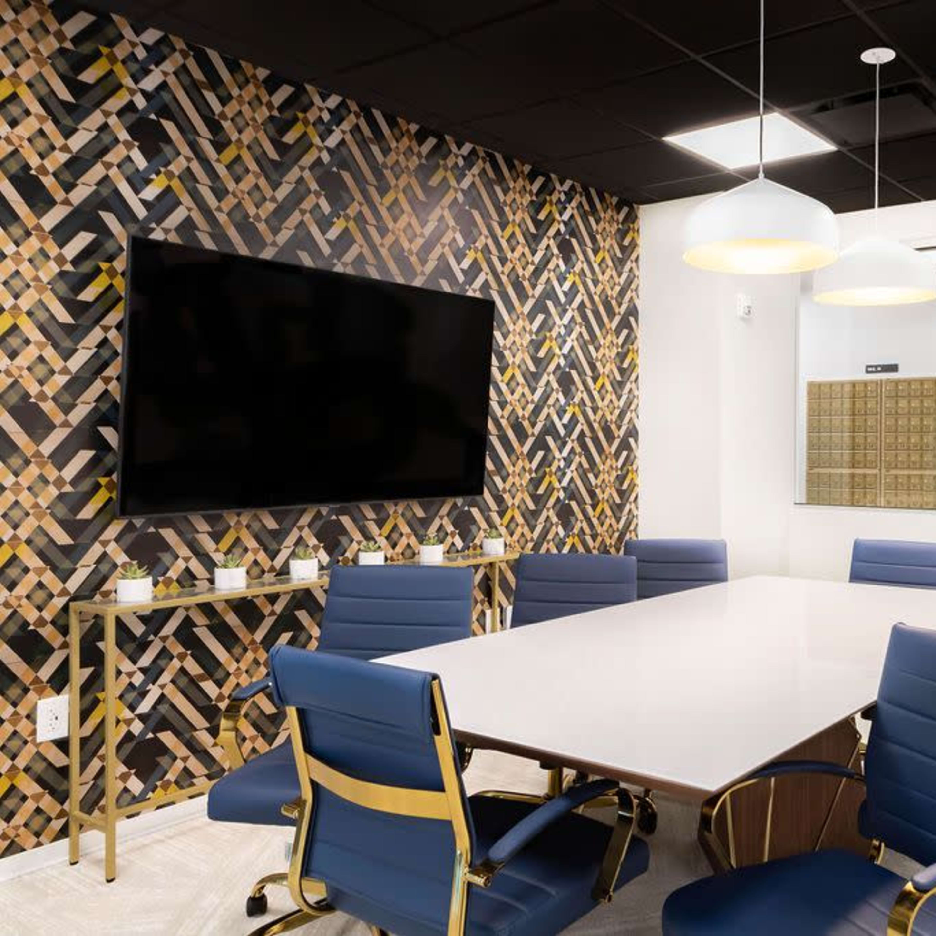 A modern conference room features a large table surrounded by blue chairs, a television on the wall, and a decorative wall with a geometric pattern.