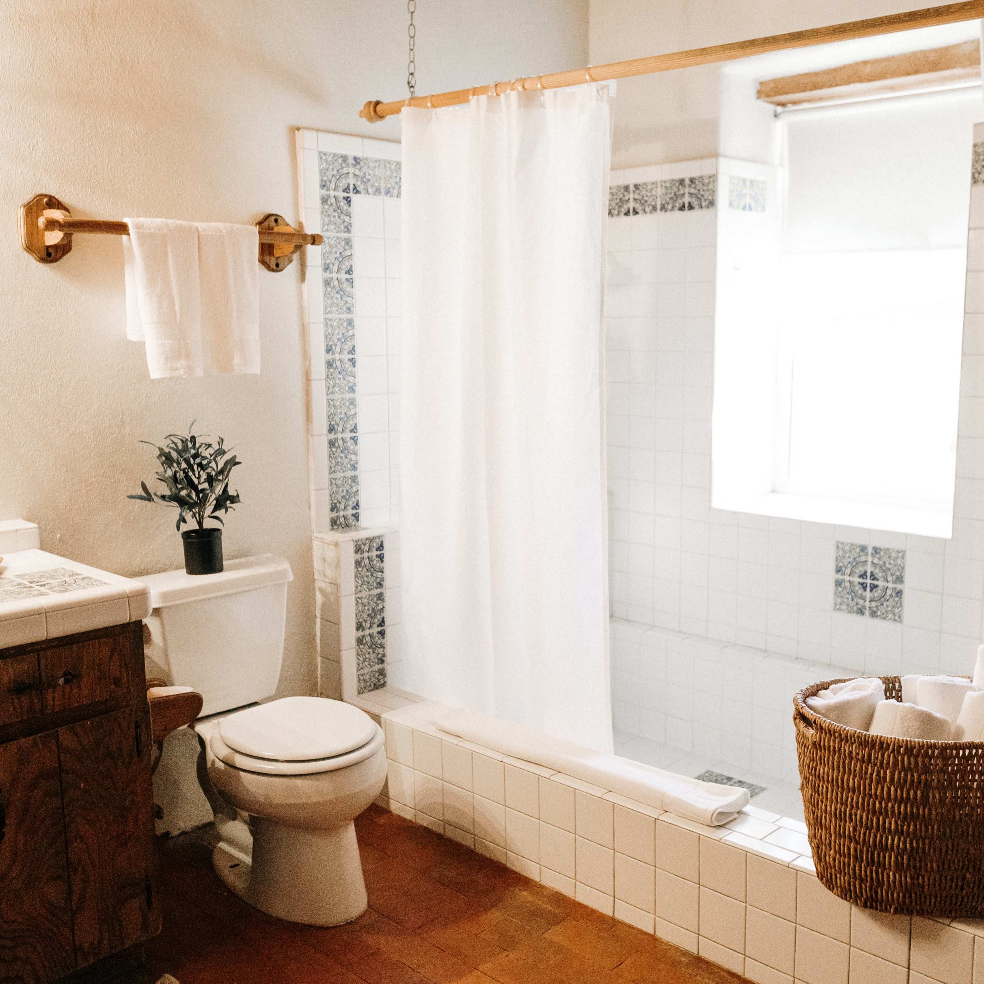 The bathroom features a shower with a white curtain, a toilet, a wooden vanity with a sink, and a wicker basket filled with rolled towels.