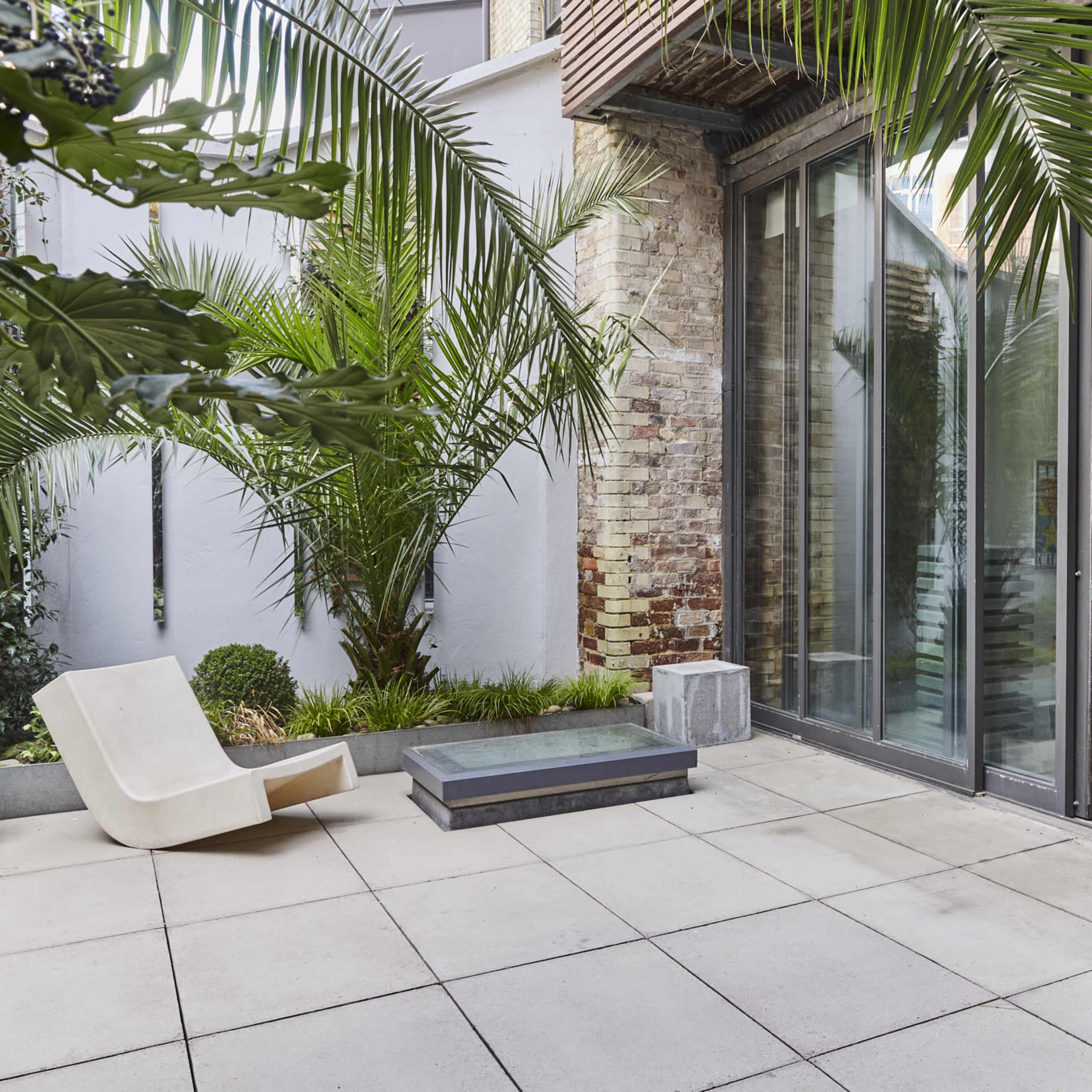 A modern outdoor space features a paved patio with a sleek lounge chair, surrounded by tropical plants and large glass doors leading into an interior area.