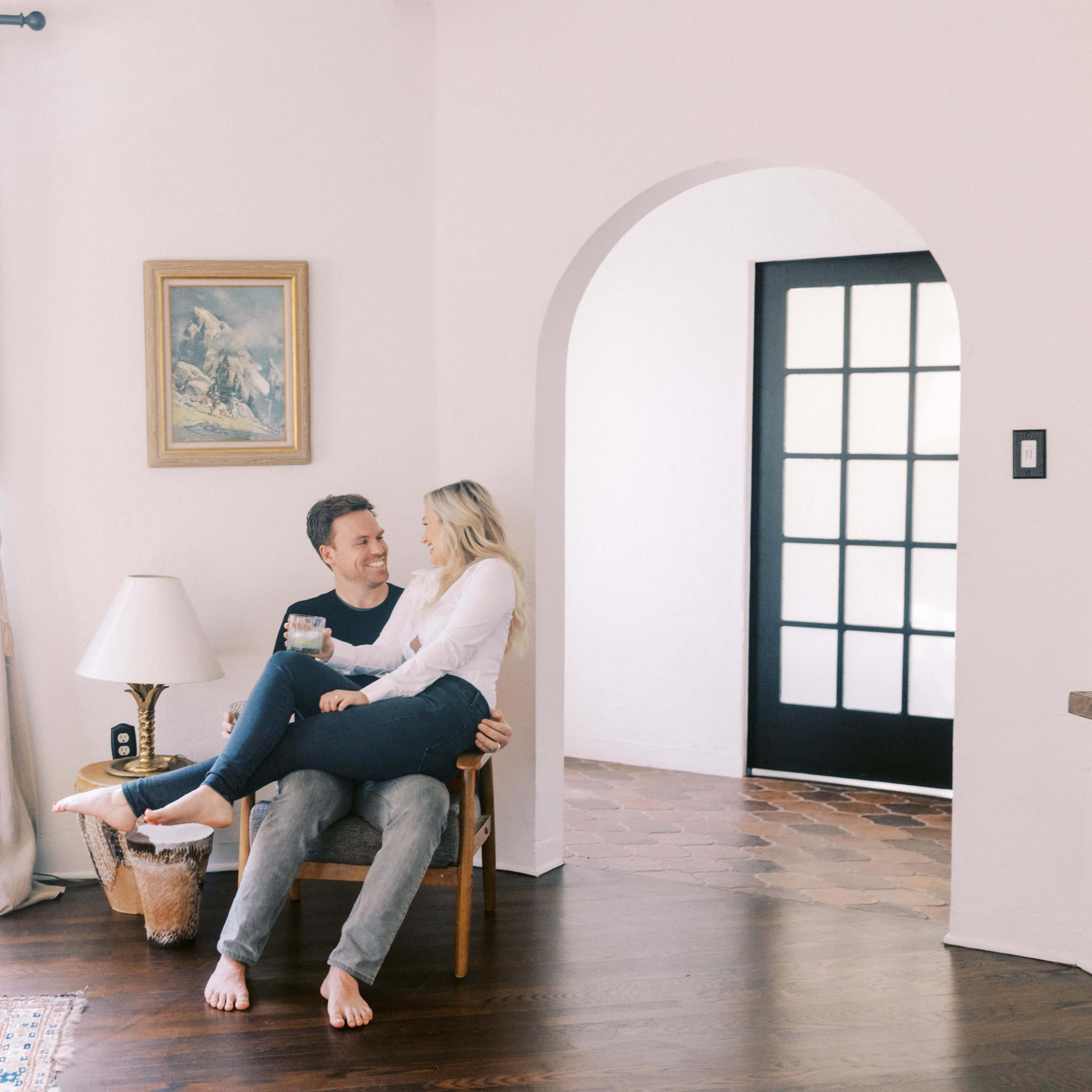 A couple sits together on a chair in a bright, airy room with large windows and an arched doorway.