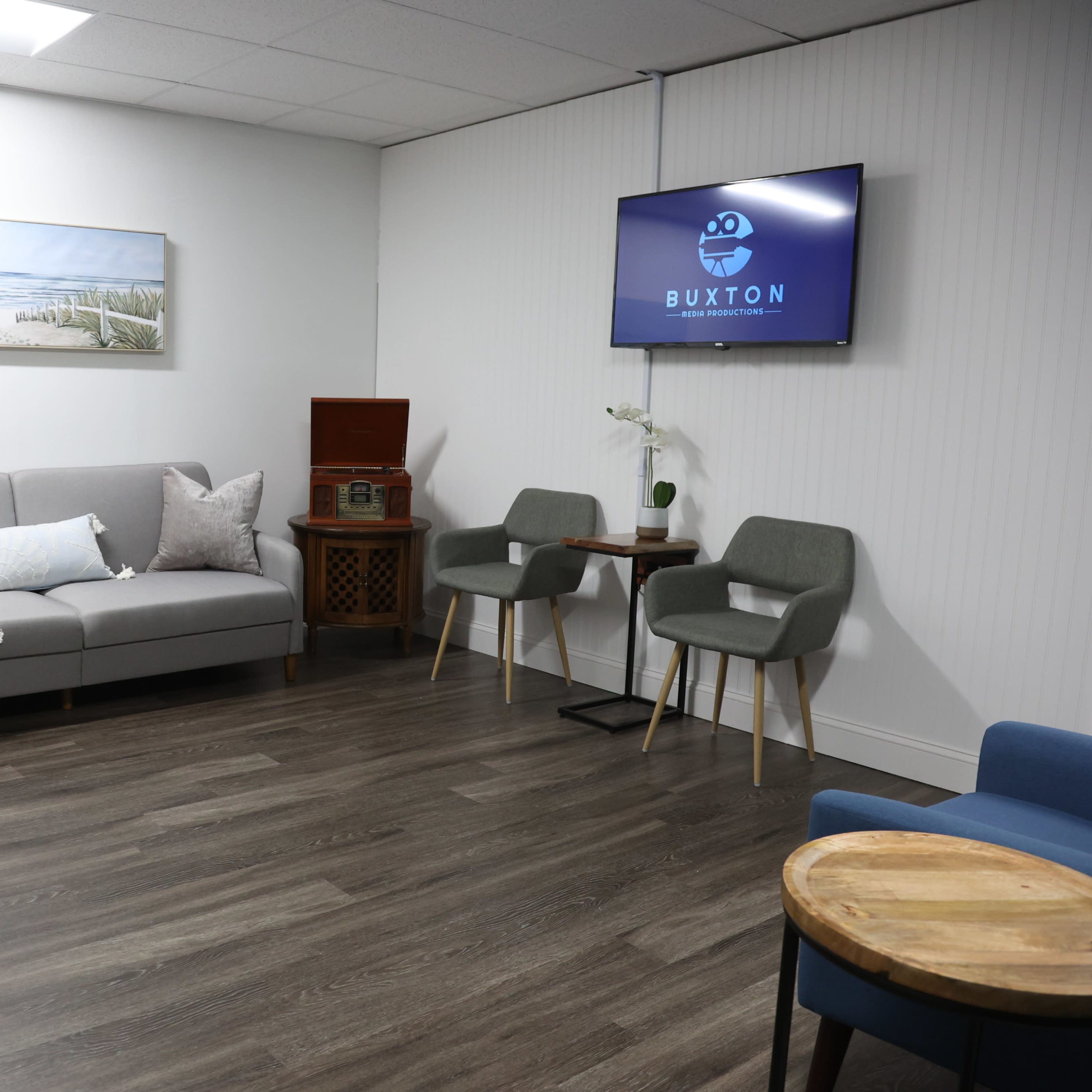 A modern waiting room featuring a sofa, two chairs, a wooden side table, an antique cabinet, and a television displaying the name "Buxton."