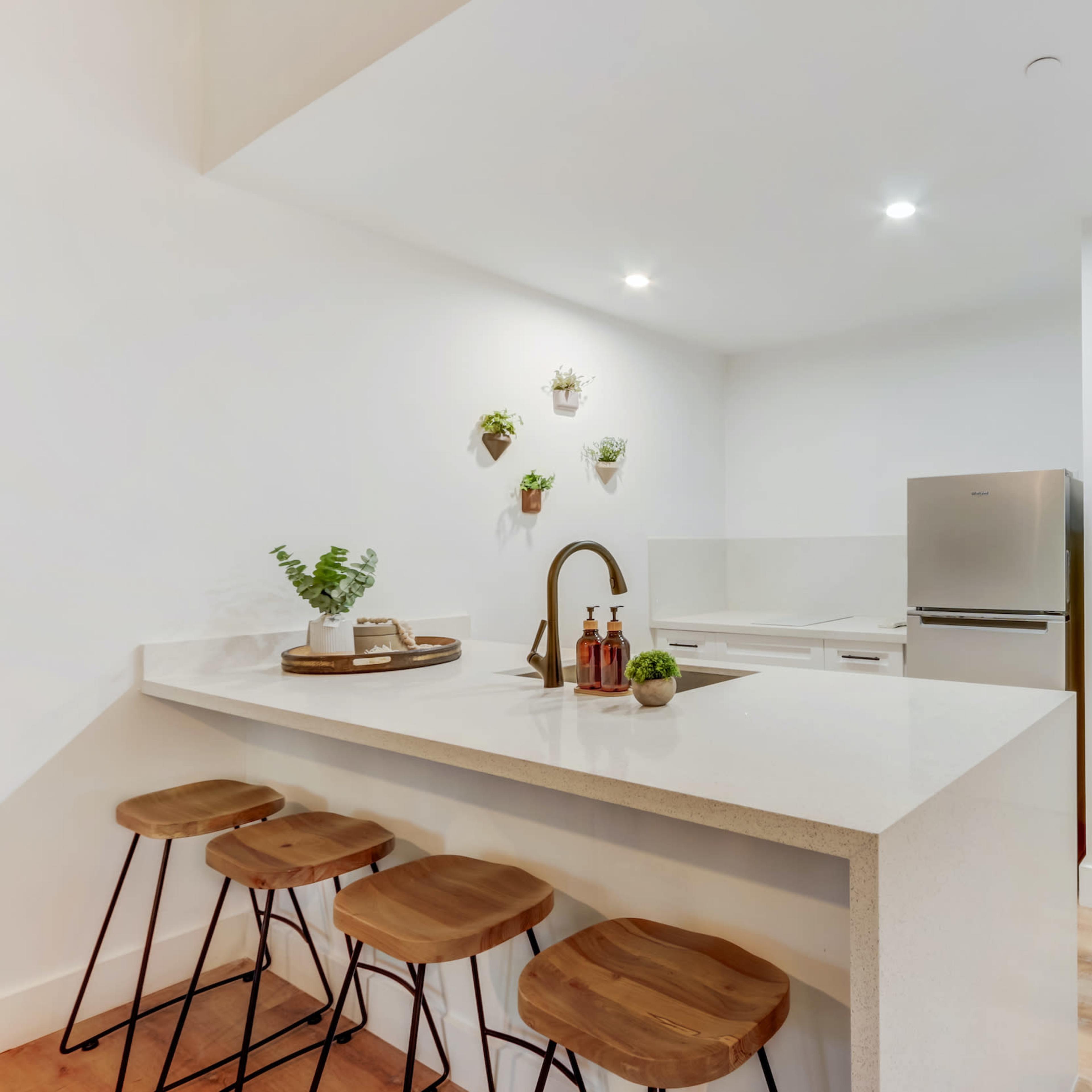 A modern kitchen with a white countertop island and three wooden stools, featuring a stainless steel refrigerator and a white door.