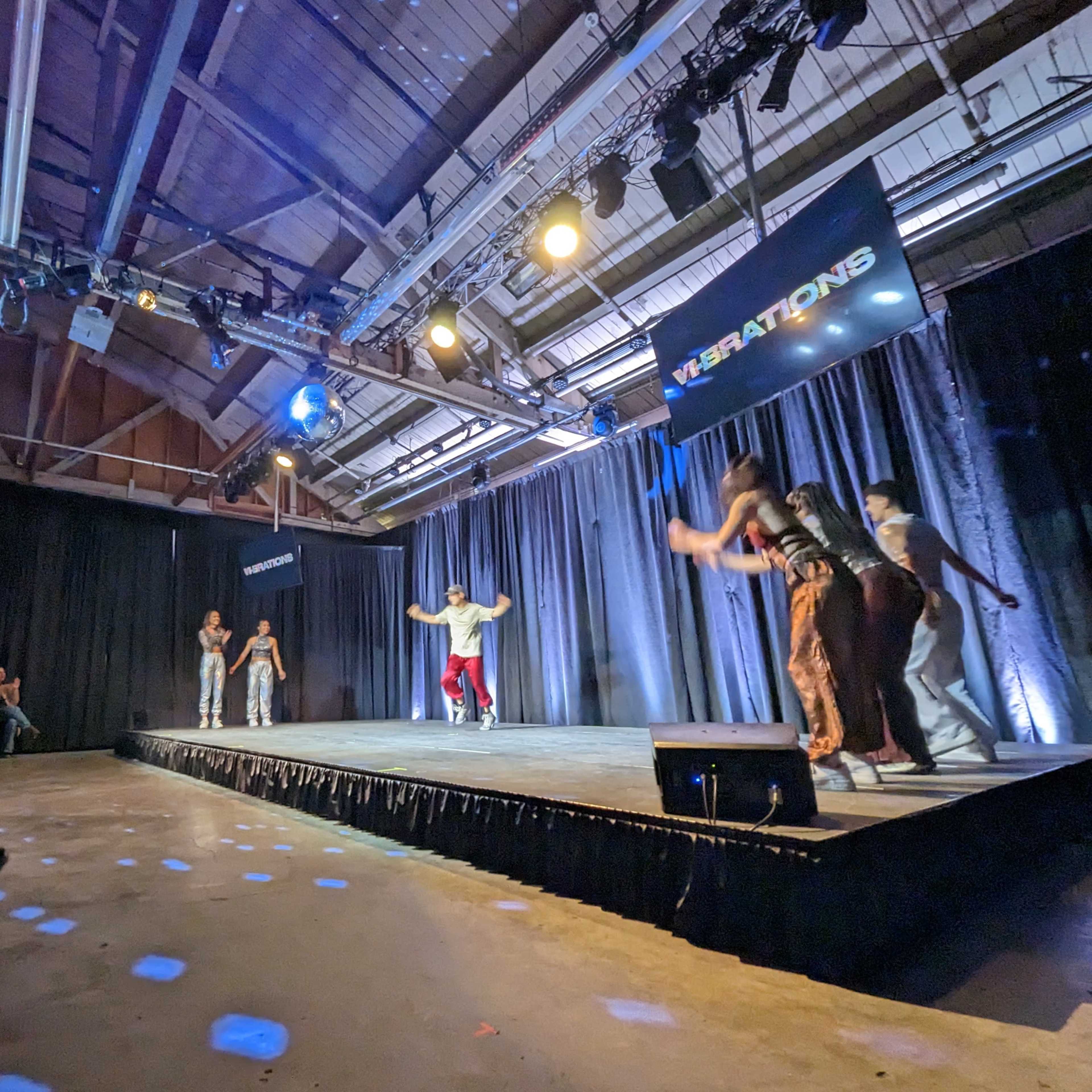 A group of dancers performs on a stage while an audience watches from the side.