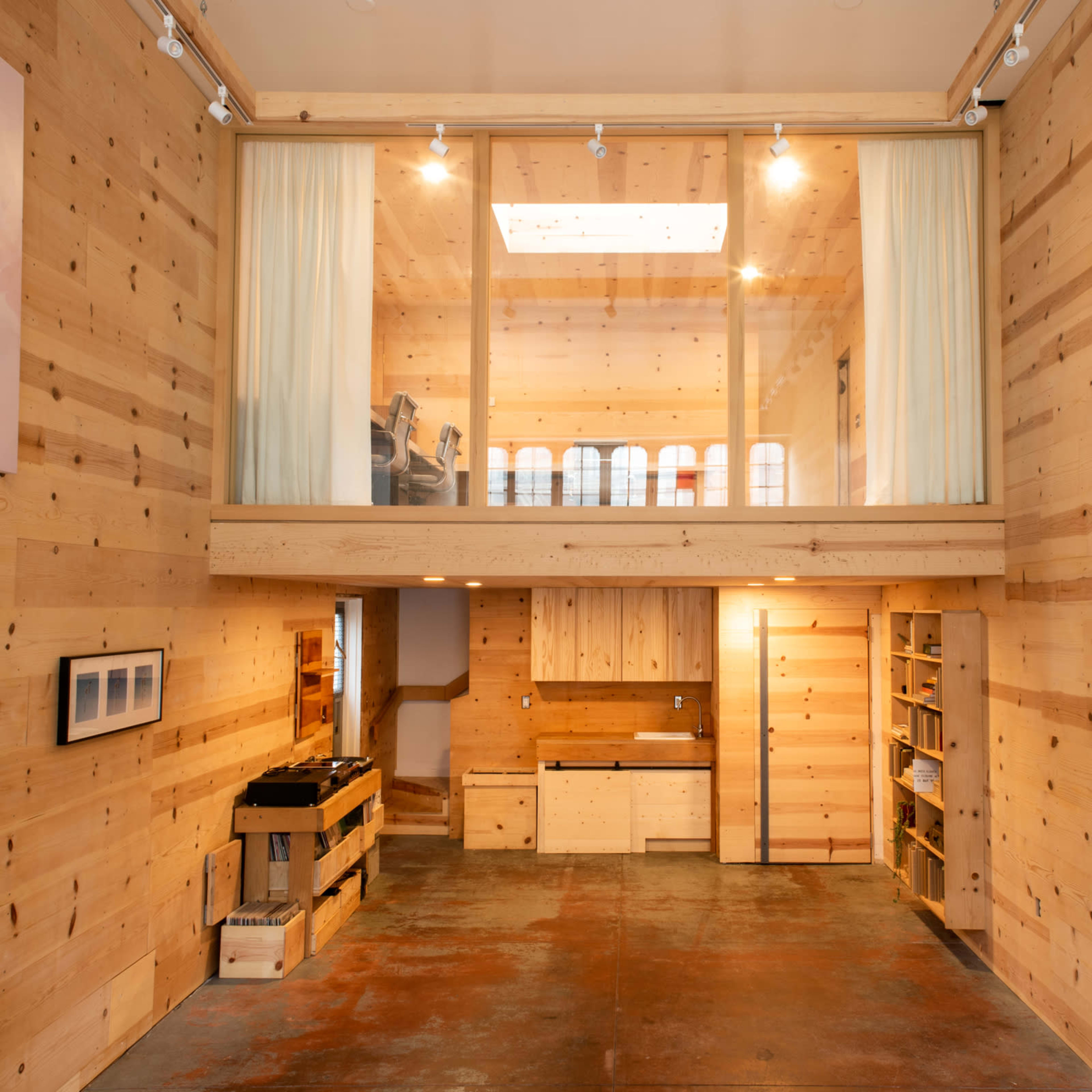 A spacious interior of a wooden room with a mezzanine level, featuring minimalist furniture, large windows, and polished concrete flooring.