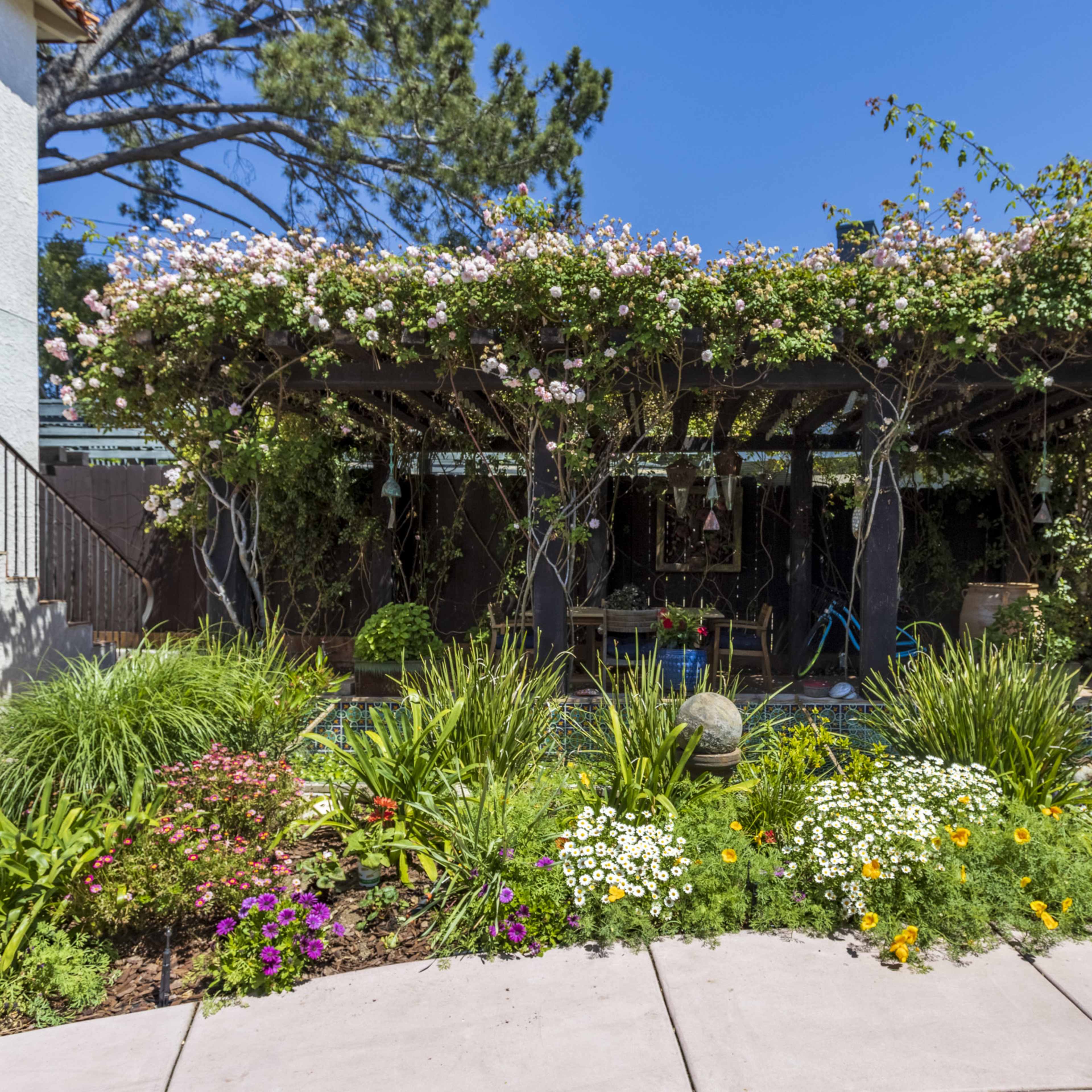 A shaded patio covered in flowering vines is surrounded by vibrant flowerbeds and green plants.