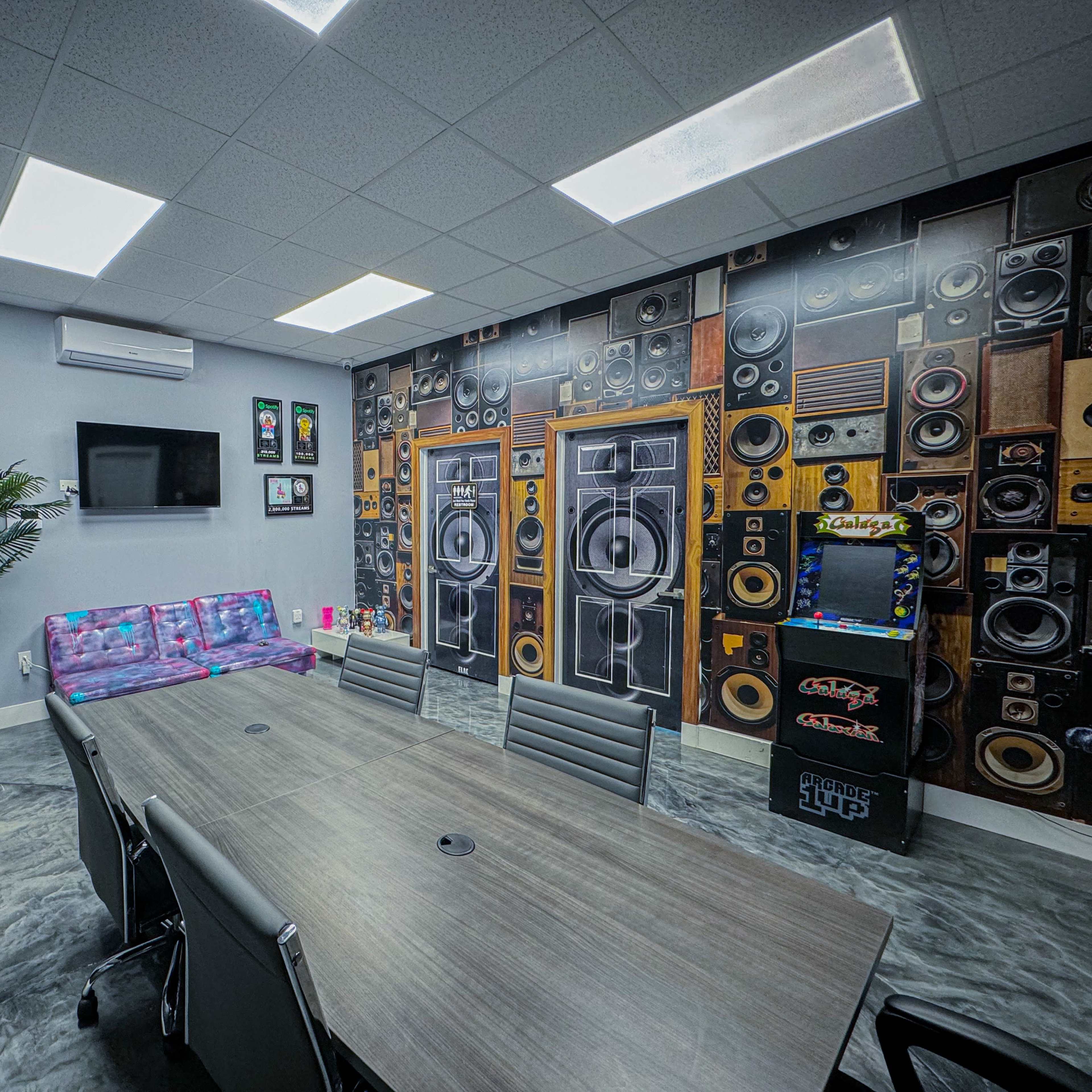 A meeting room features a long table surrounded by chairs, a wall decorated with large speaker graphics, and a couch along one side.