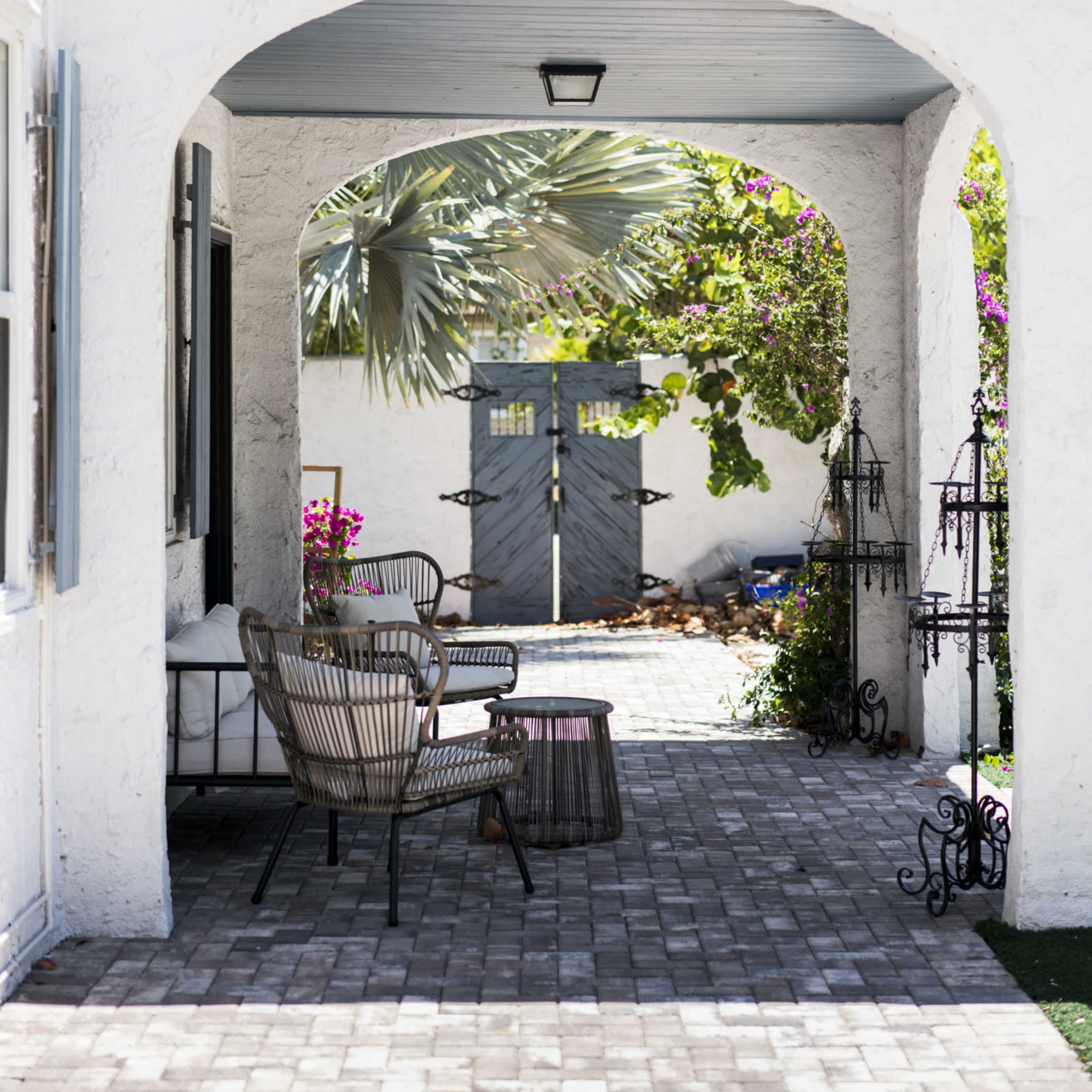 A shaded outdoor passage features a paved walkway, two chairs, and a gated entrance lined with greenery.