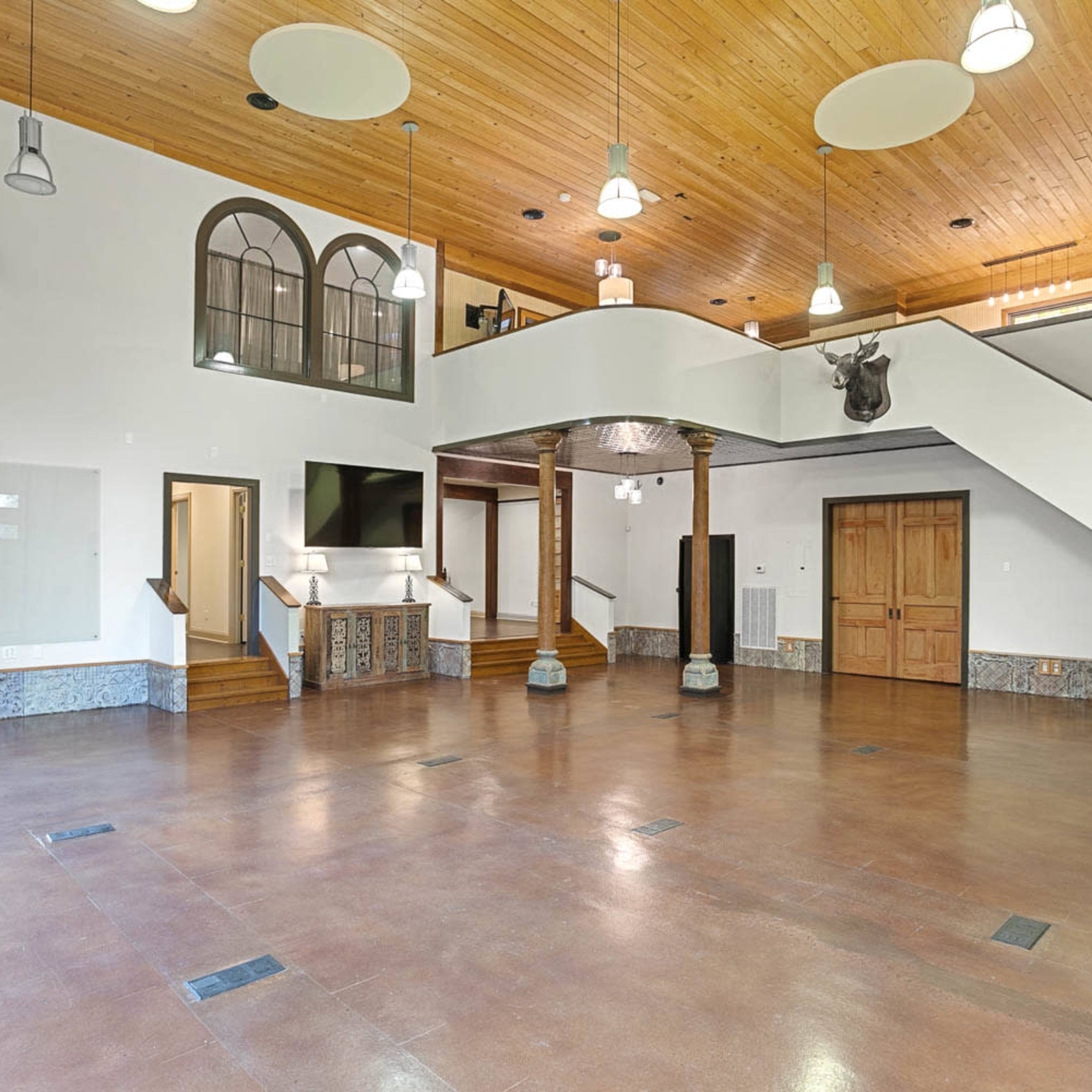 A spacious interior of a building with wooden ceiling, large windows, and a staircase leading to a second level.