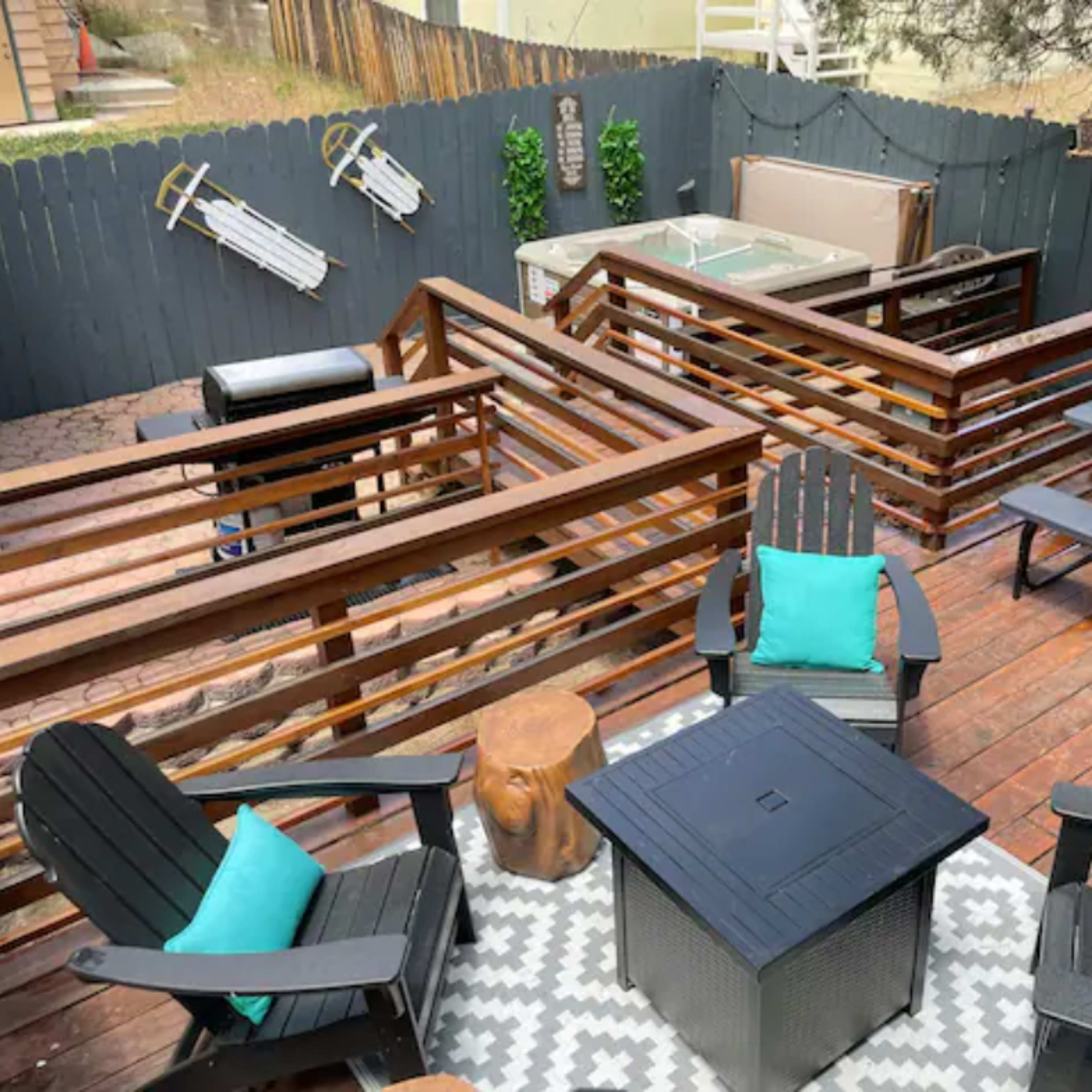 A wooden deck area with black chairs, a square table, and a hot tub, surrounded by dark wooden railings and greenery.