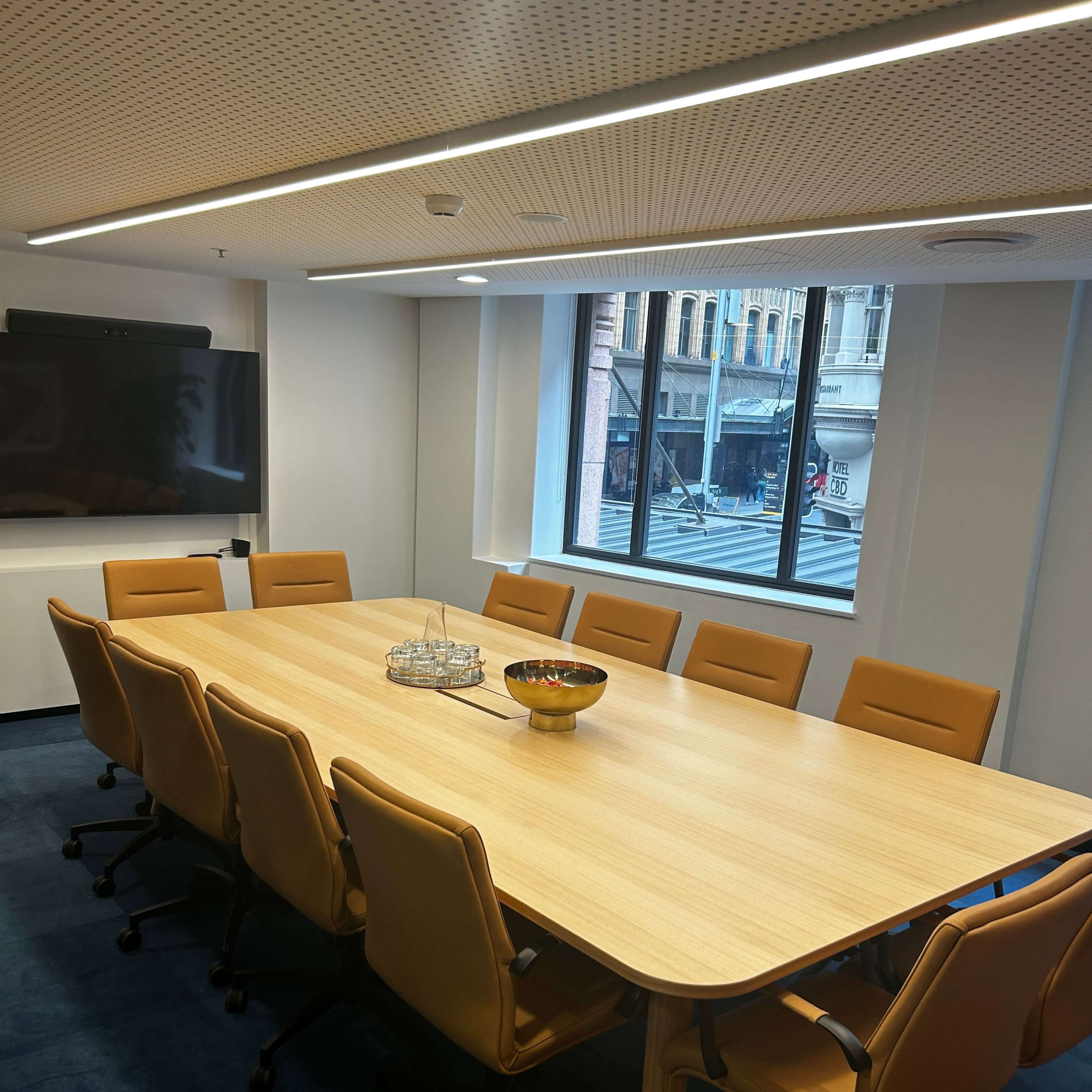 A large conference room features a wooden table surrounded by ten orange chairs, with a bowl of fruit in the center and a view of the street through a window.