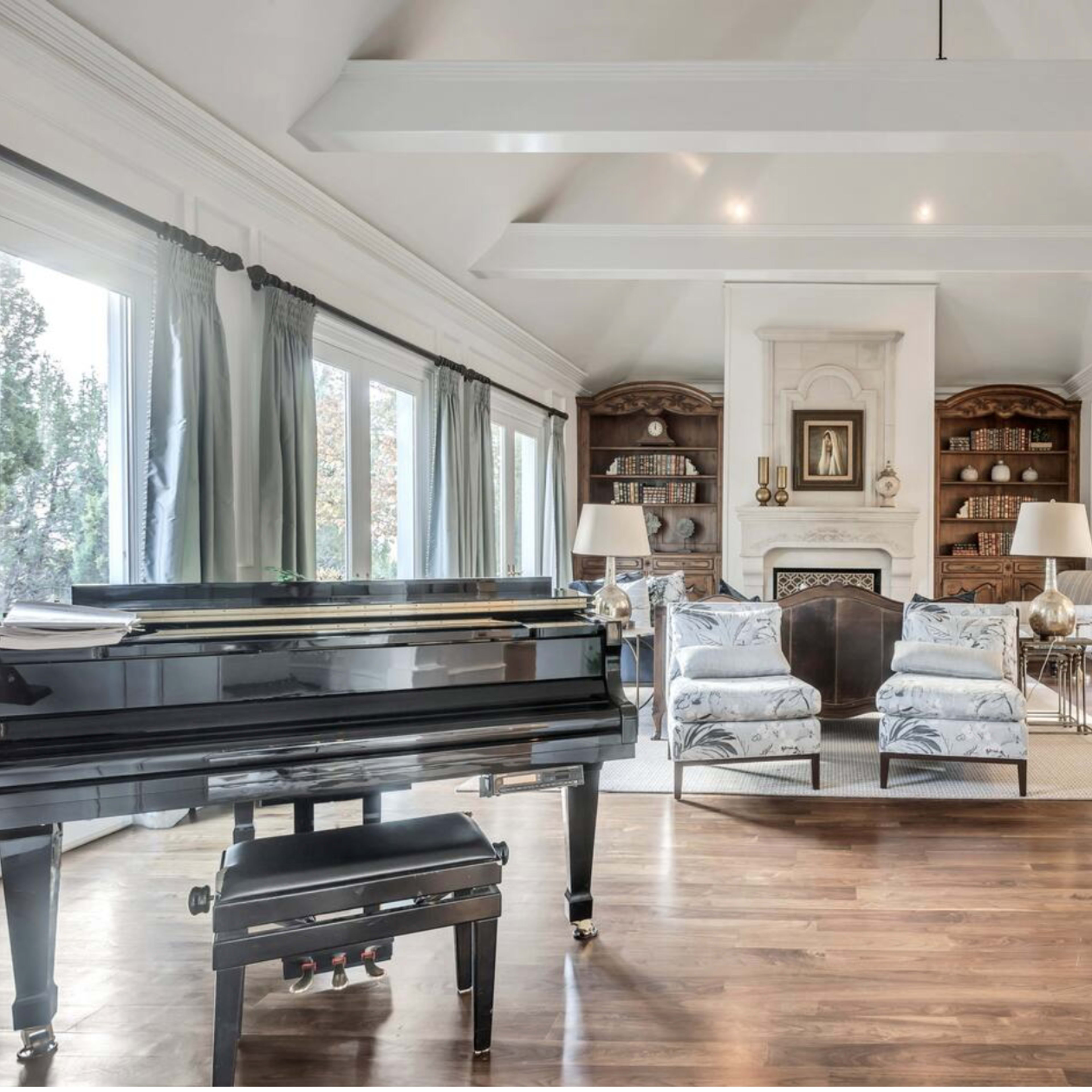 A spacious living area featuring a grand piano, two patterned armchairs, built-in bookshelves, and a dining area in the background.