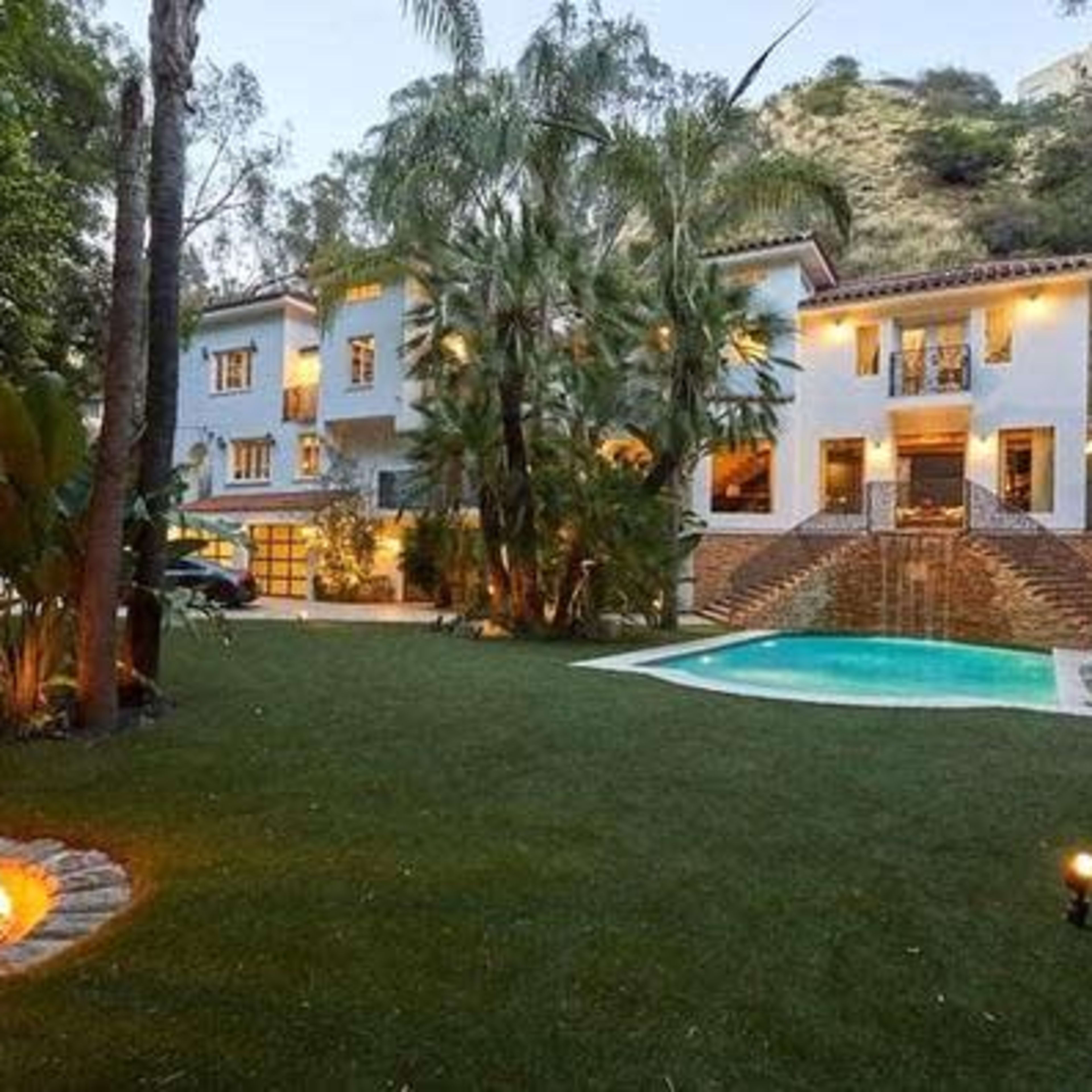 A two-story house surrounded by palm trees and a manicured lawn, featuring a pool and a fire pit in the backyard.