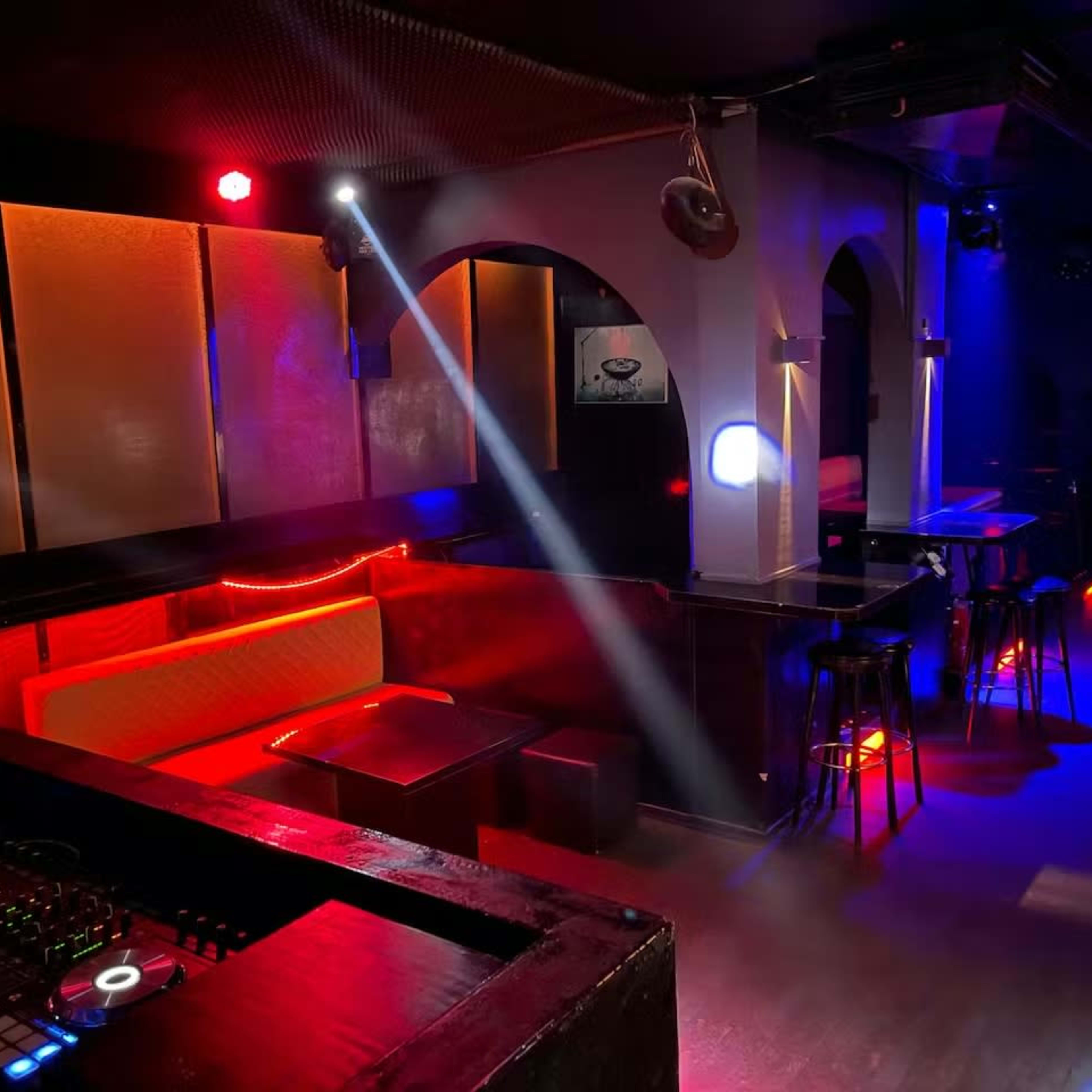 A dimly lit bar or nightclub interior with illuminated seating areas and electronic equipment at the foreground.
