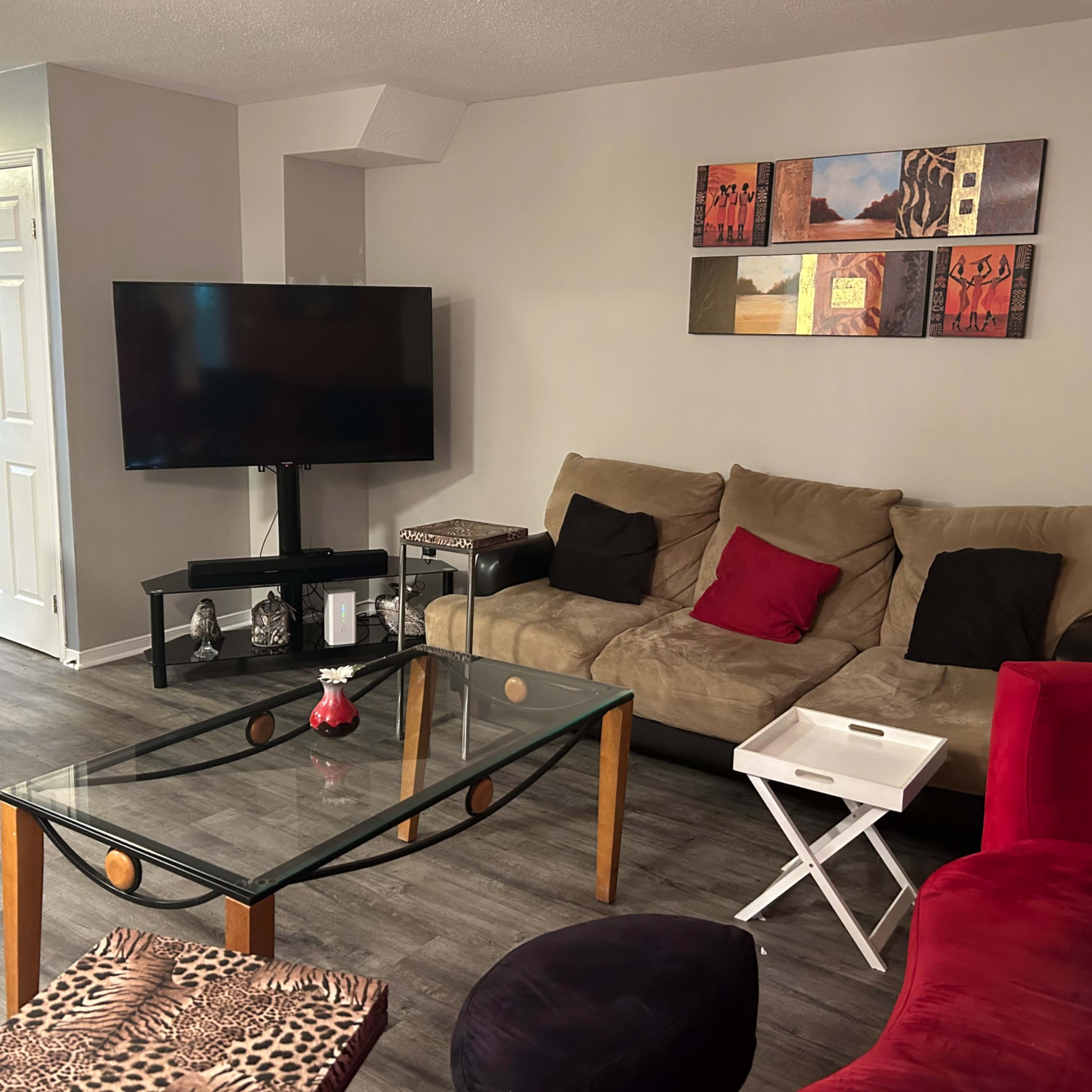 The living room features a beige sofa with red and black accents, a glass coffee table, and a large television set against a neutral wall.