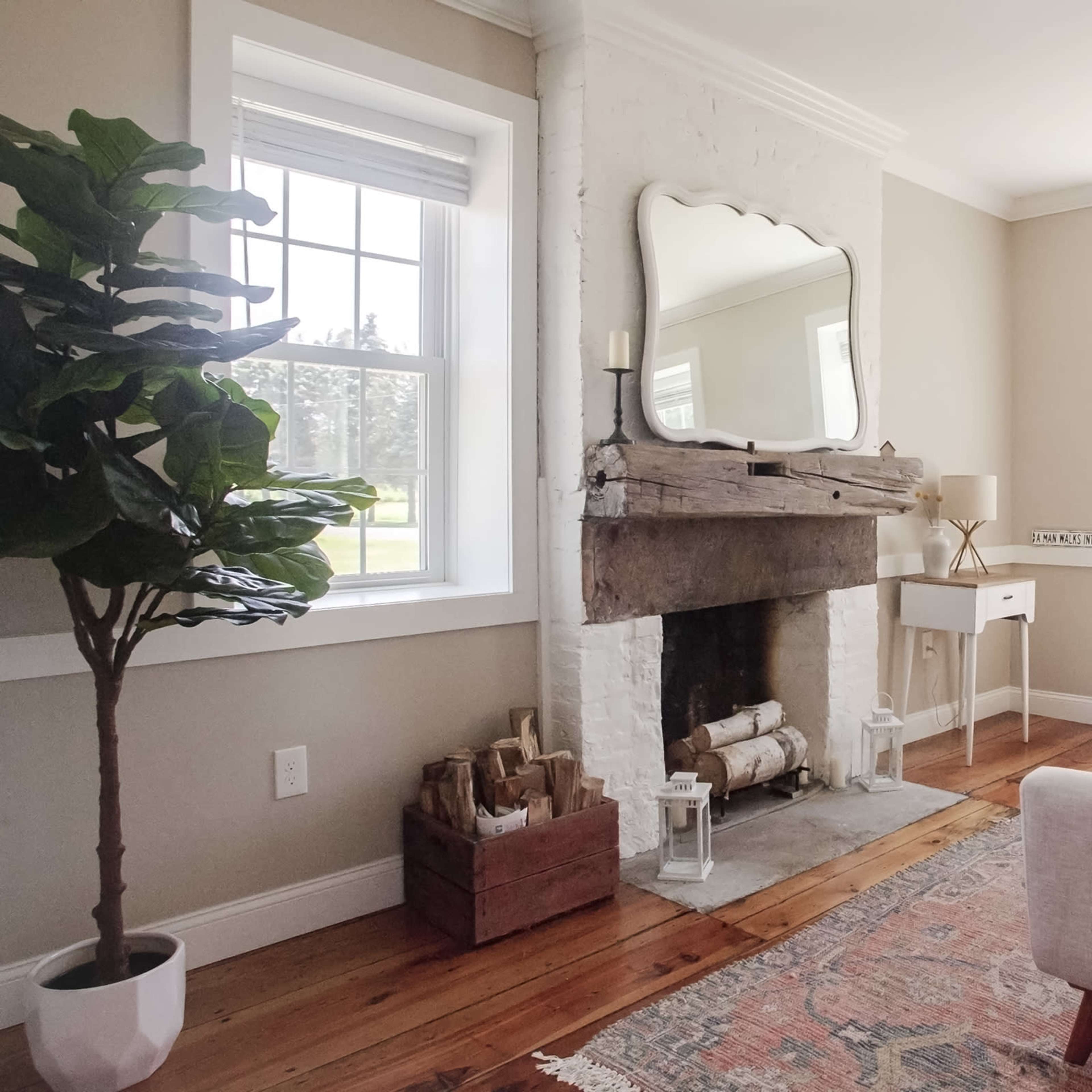 A cozy living room features a white brick fireplace with a wooden mantel, a large mirror above it, and a sofa facing two windows.