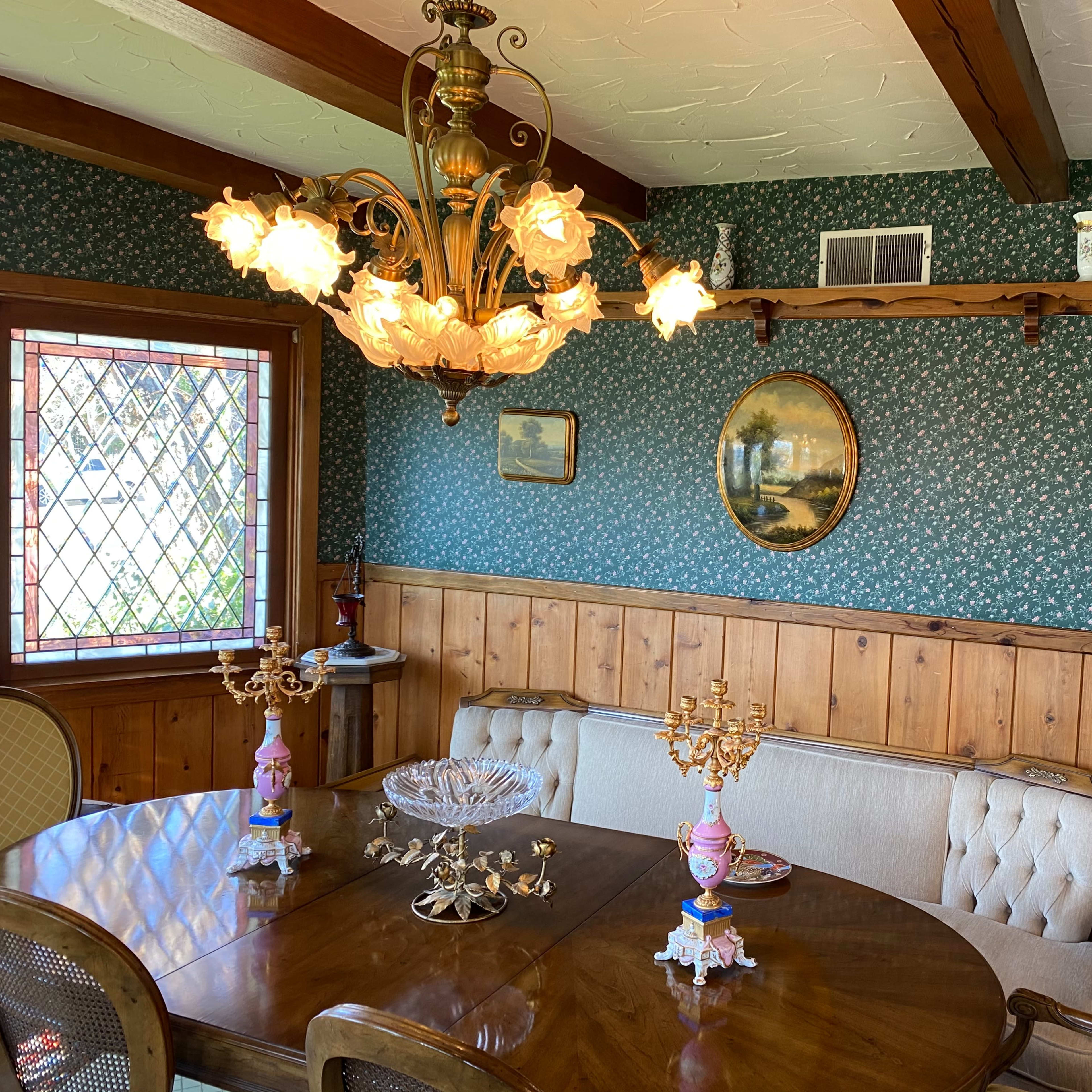 A dining room featuring a wooden table surrounded by upholstered chairs, a chandelier with floral shades hanging above, and floral wallpaper with framed paintings on the walls.