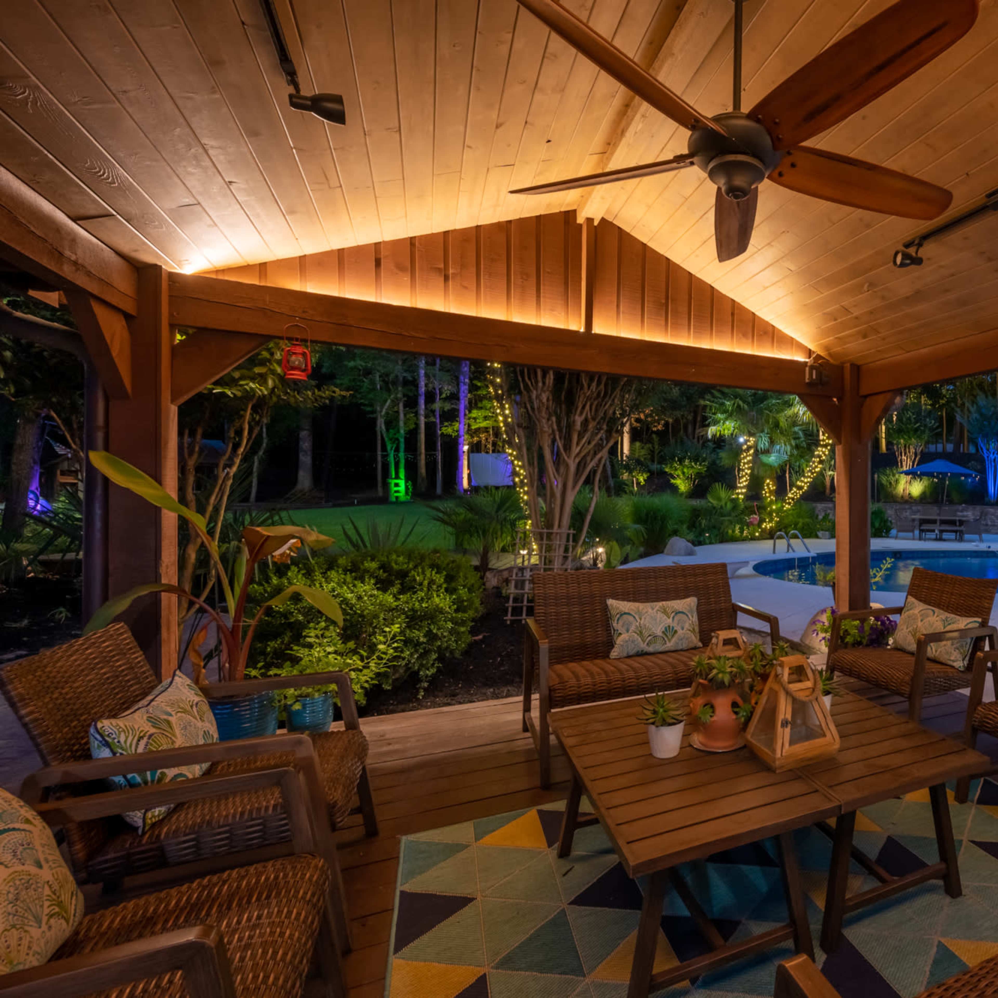 A covered outdoor seating area features wicker chairs and a wooden table, surrounded by a well-lit garden and a pool area.