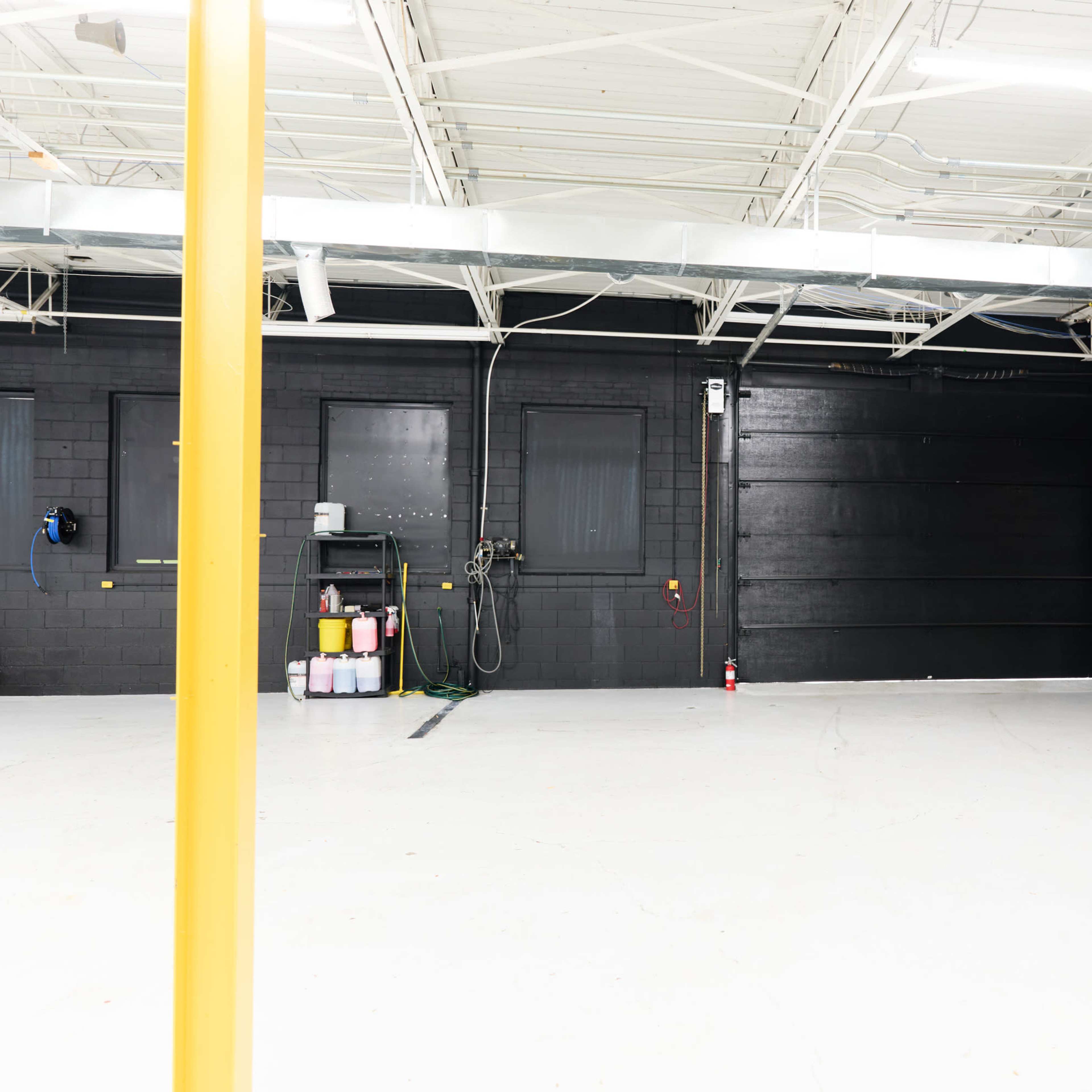 An empty industrial workspace with a polished white floor and black walls, featuring a yellow pole on the left and various equipment along the back wall.