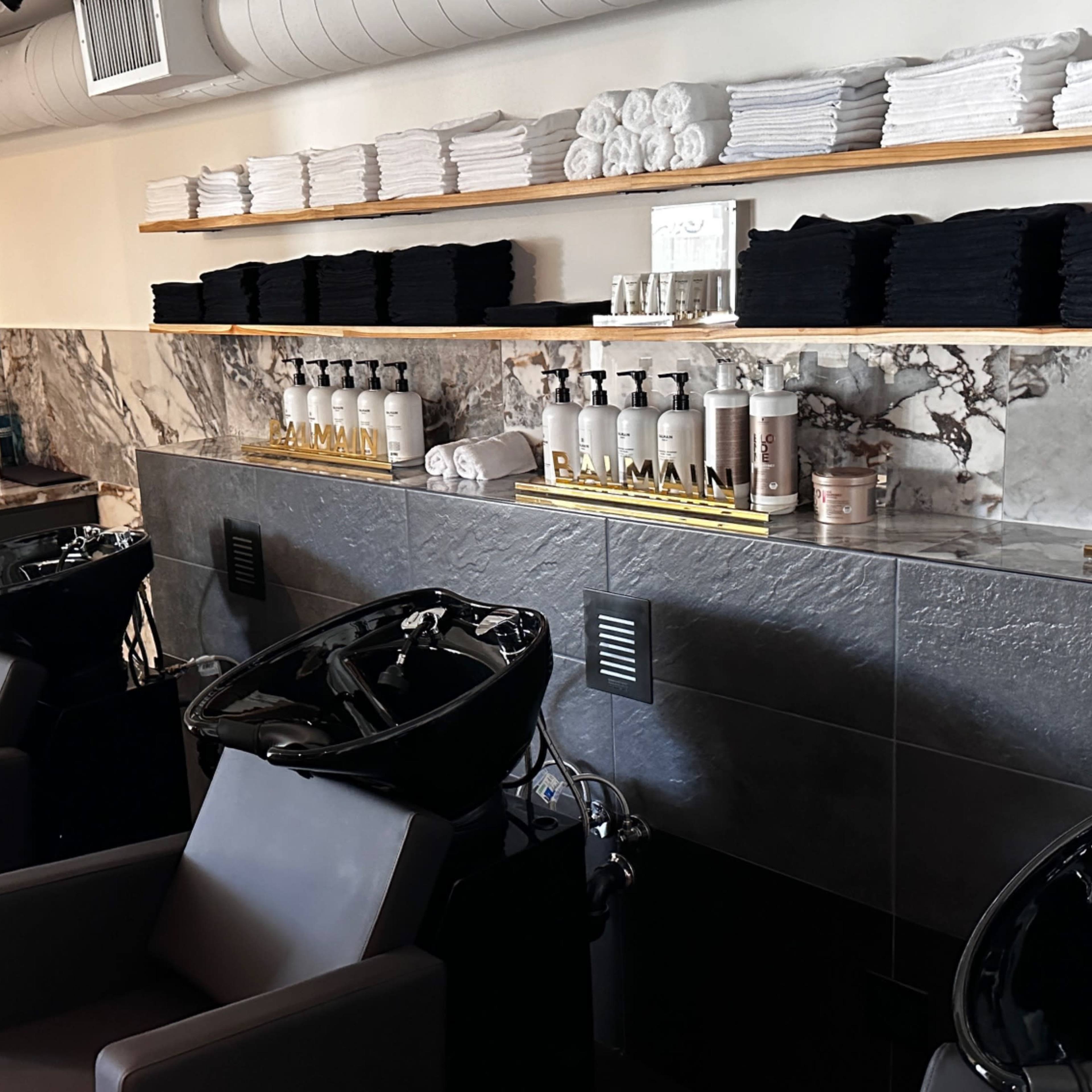 A modern salon area with black shampoo chairs, a marble counter, and neatly stacked towels on shelves.