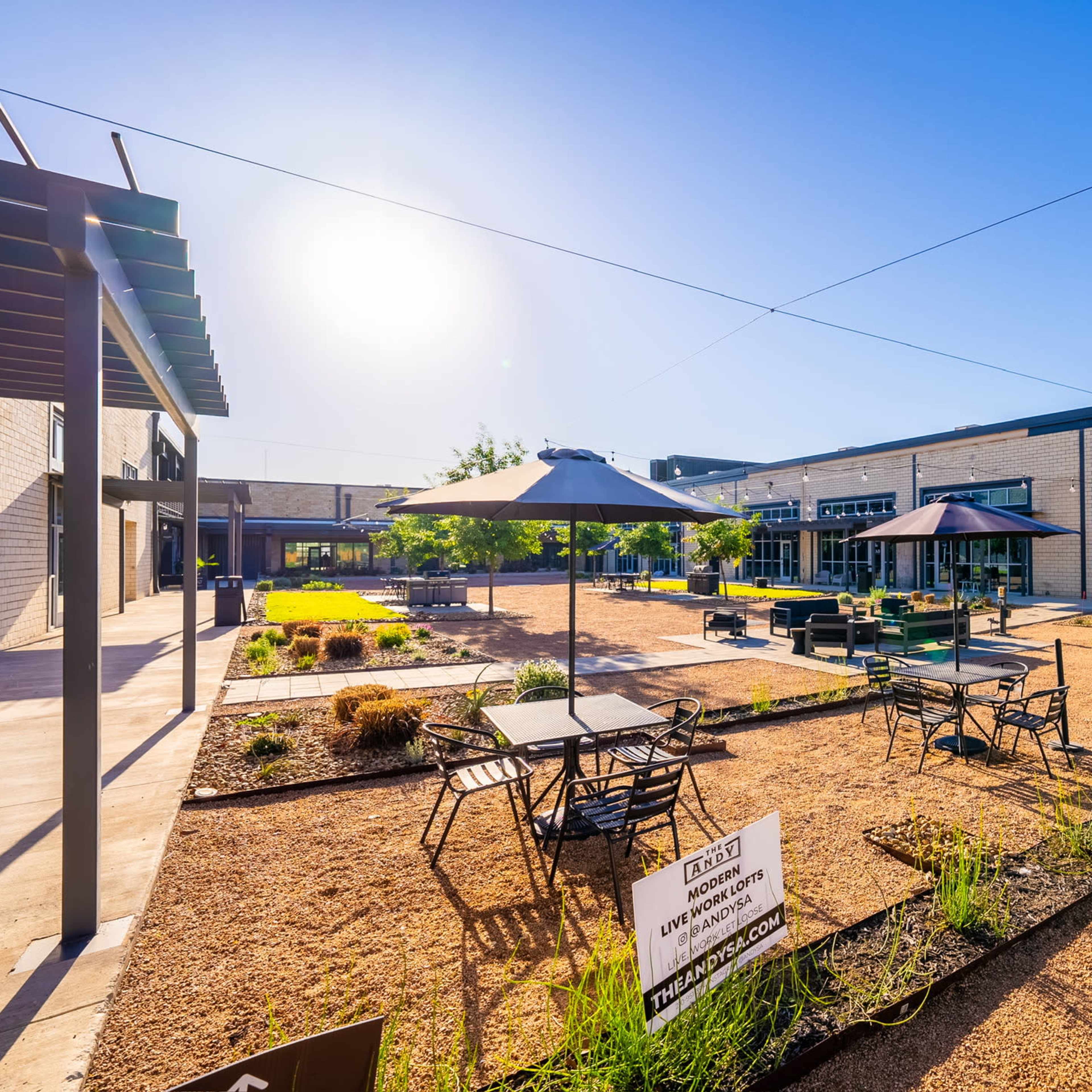 A landscaped courtyard with tables and umbrellas surrounded by modern buildings, featuring greenery and gravel pathways under clear blue skies.