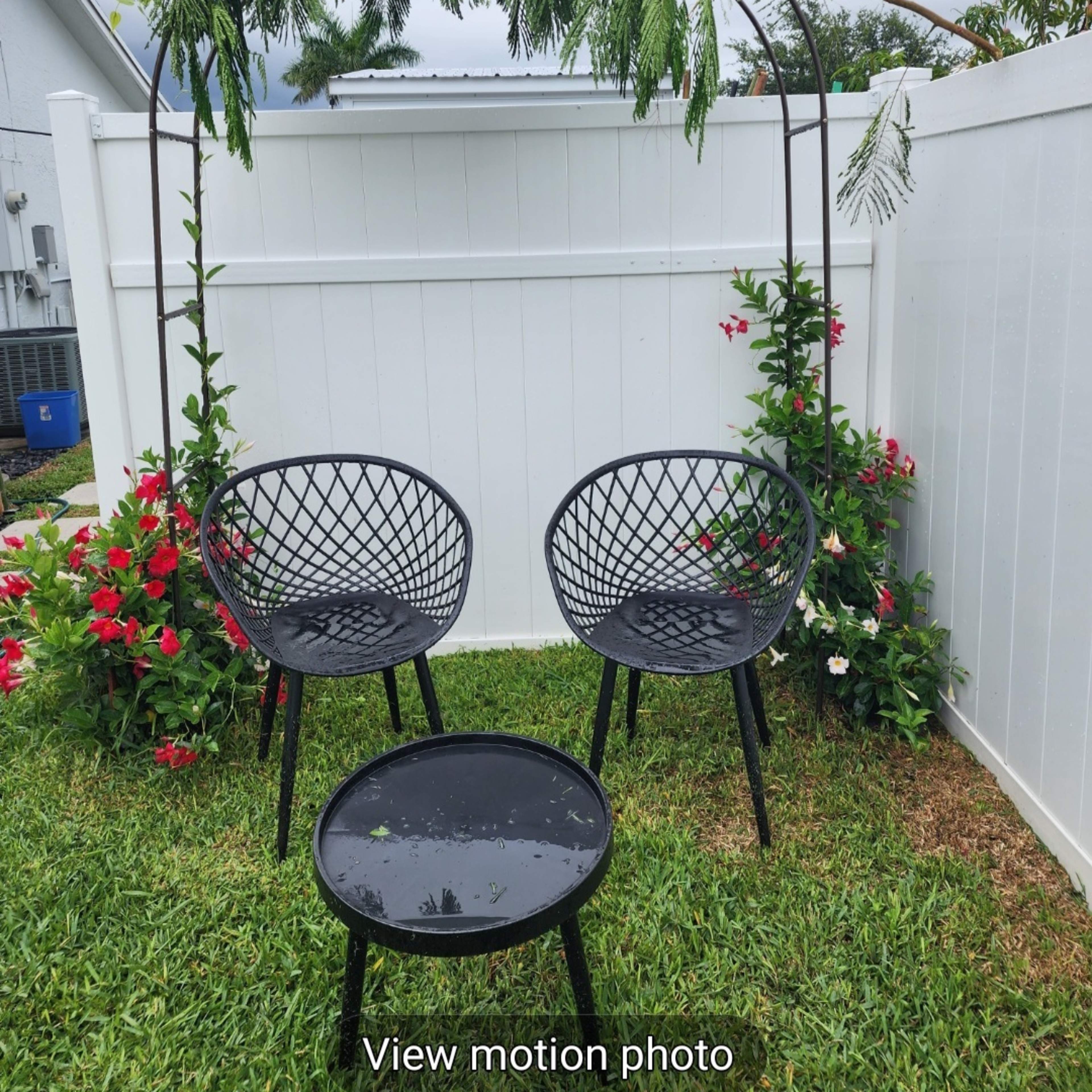 A decorative arch adorned with greenery and white flowers stands over three black wire chairs and a small table in a garden setting.